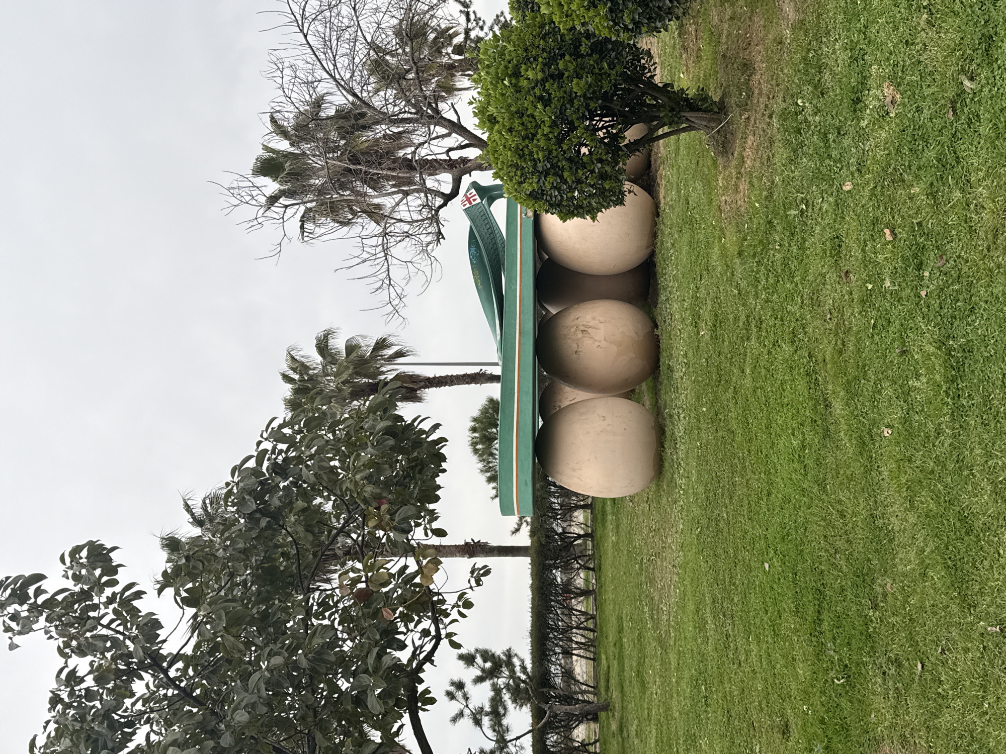 The image depicts a large, green flip-flop sculpture resting on four spherical stones in a grassy area. The location is likely Batumi, Georgia, as a British flag is visible on the sandal's strap. Vegetation surrounds the sculpture, including trees, bushes, and green grass. The sky is overcast, suggesting a cloudy day. The scene is devoid of people, focusing on the artwork and its immediate environment.