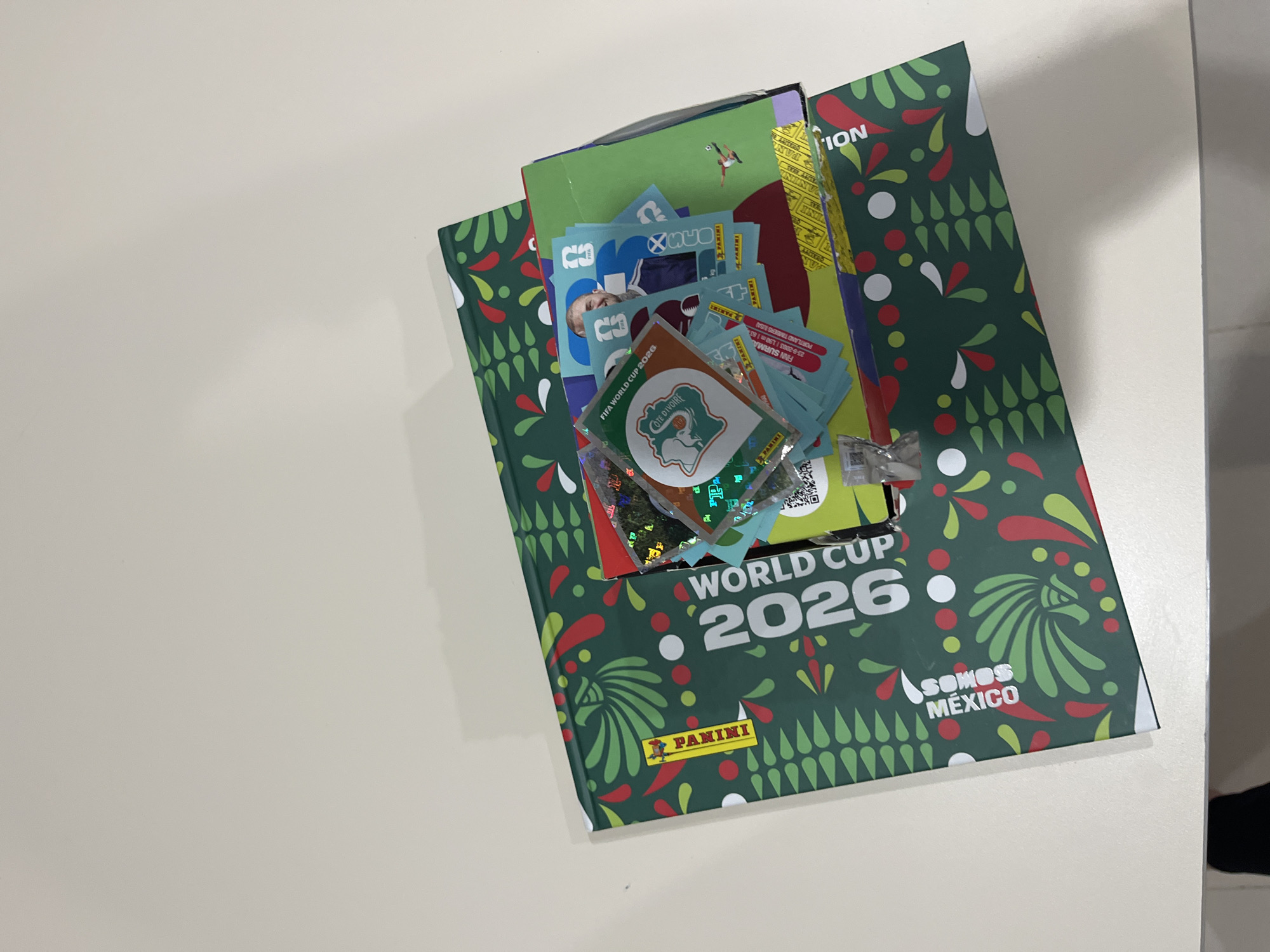 The image shows a Panini FIFA World Cup 2026 sticker album. The album is green and decorated with red, green, and white patterns. "WORLD CUP 2026" is printed in white lettering. There is also a "PANINI" logo and the words "SOMOS MEXICO". The album is open, with a stack of sticker packs and individual stickers placed on top. A sticker for Cote d'Ivoire is visible on top of the pile. The album is on a light-colored surface. There is no indication of San Bernardino Tlaxcalancingo, Mexico.