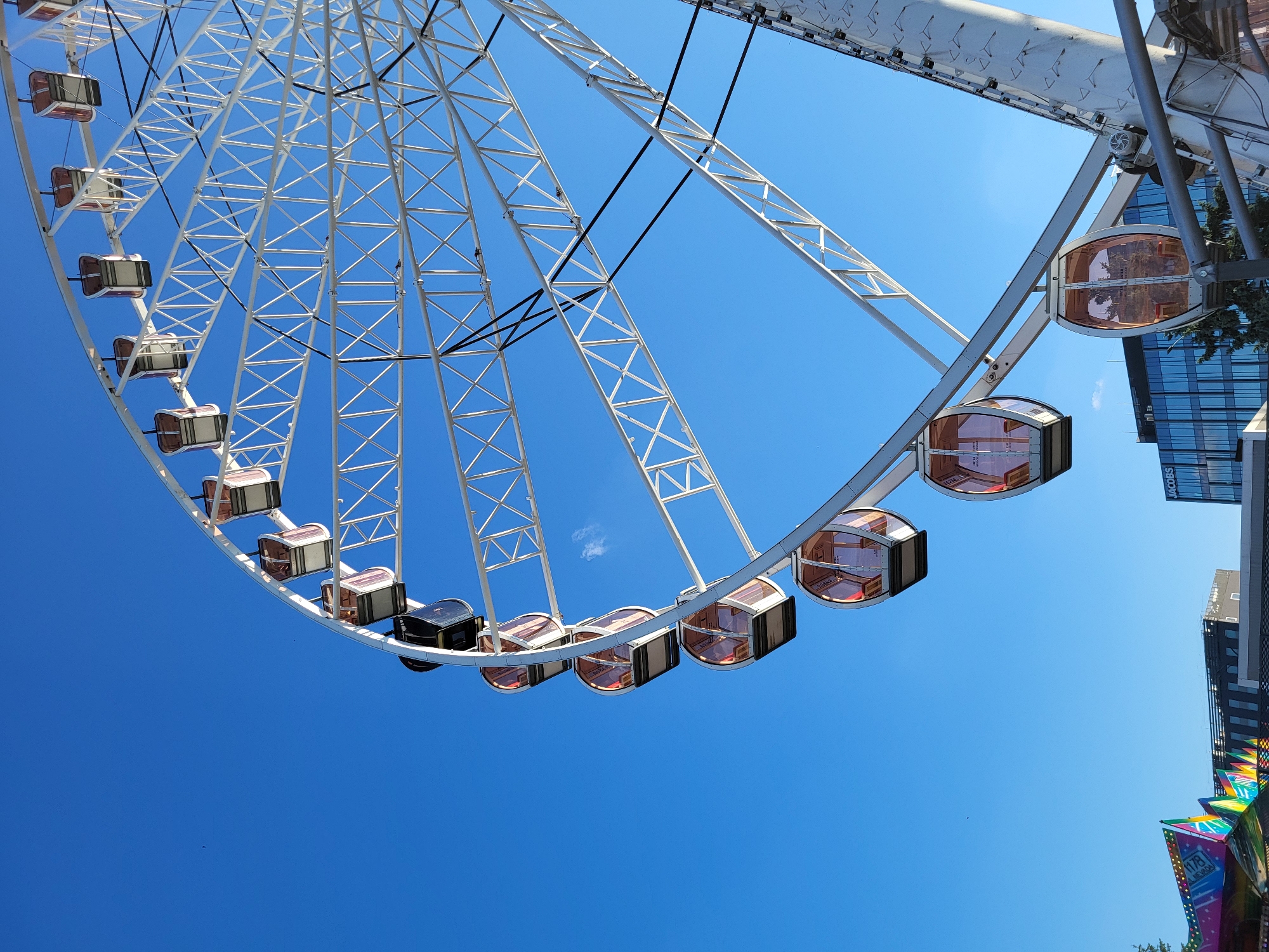 The image captures a detailed view of a large Ferris wheel set against a clear, deep blue sky. The Ferris wheel, constructed with a white metallic frame, occupies the right and upper portions of the frame, showcasing its intricate lattice of spokes and structural supports. Multiple enclosed passenger gondolas, featuring amber-tinted windows and dark upper frames, are suspended from the wheel's circumference, some appearing empty. One gondola near the top is notably dark-colored.
In the background, beneath the Ferris wheel and towards the right, modern city architecture is visible, including a multi-story building with dark facades and numerous reflective glass windows. A prominent white sign displaying "JACOBS" is affixed to the upper section of this building. Below the "JACOBS" building, a patch of green foliage is discernible. In the bottom-left corner of the image, a section of another brightly colored amusement park ride, featuring panels in hues of yellow, red, blue, and green, is partially visible. The sky, a dominant feature, is largely cloudless, save for a faint, wispy white cloud fragment in the mid-left quadrant. Several small, dark specks, possibly distant birds, are also visible in the upper left sky. The scene is brightly lit by natural sunlight, indicating daytime. The specific location within the city of Unknown, Unknown cannot be further identified beyond the visible "JACOBS" signage on a background building.