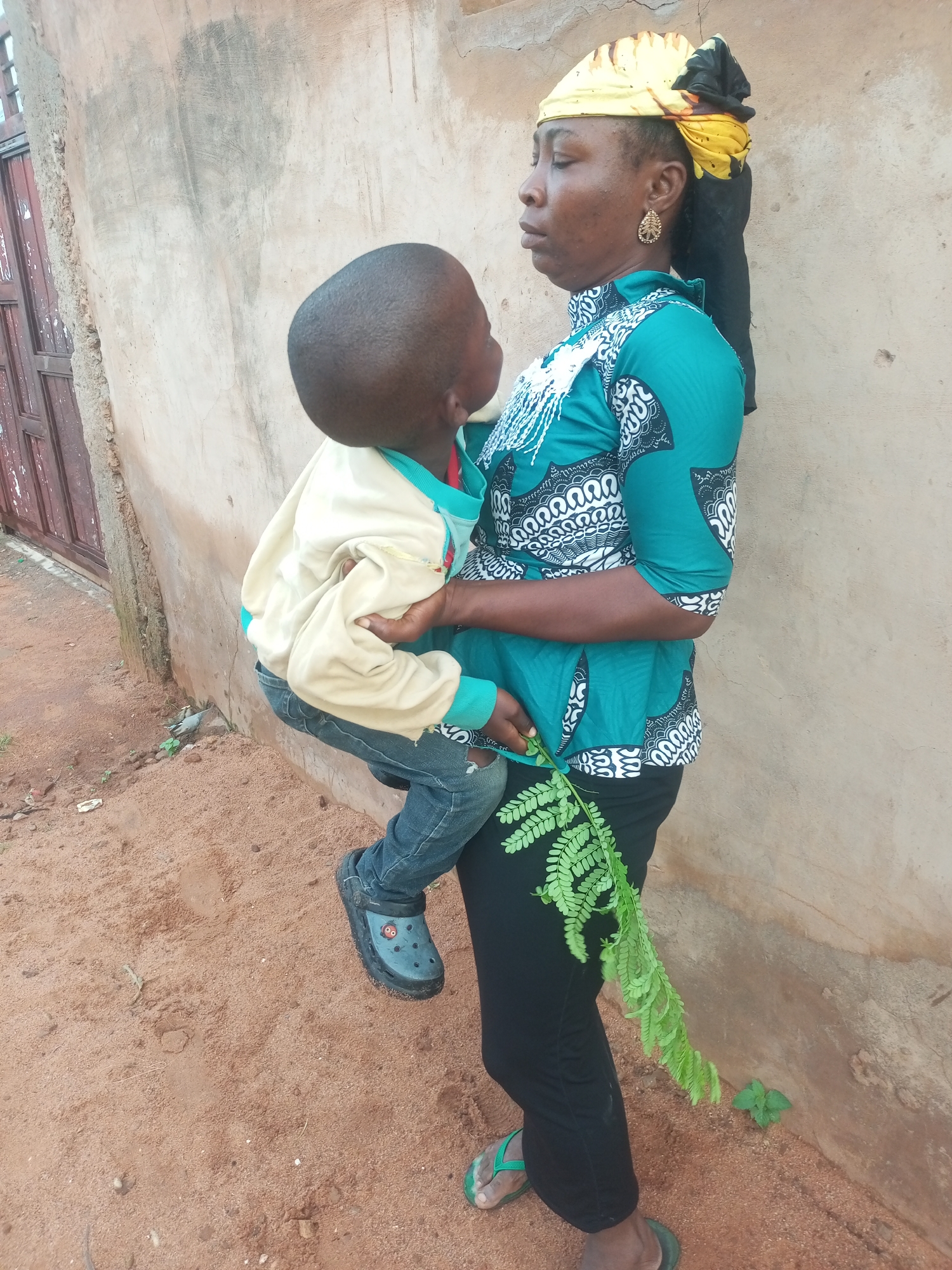 A woman is standing next to a textured, light brown wall in the city of Tempure, Nigeria. She is holding a young child, who is facing away from the viewer. The woman is wearing a green and white patterned top, black pants, and a yellow and black headscarf. She also has a large, decorative earring in her left ear. A sprig of green leaves is held in her right hand, dangling near her leg. The child is wearing a beige jacket with a teal collar and blue jeans. The ground is covered in reddish-brown dirt, with some small pebbles and a few sparse green plants. To the left of the image, a weathered wooden door is visible, leaning against the wall. The overall lighting suggests it is daytime.