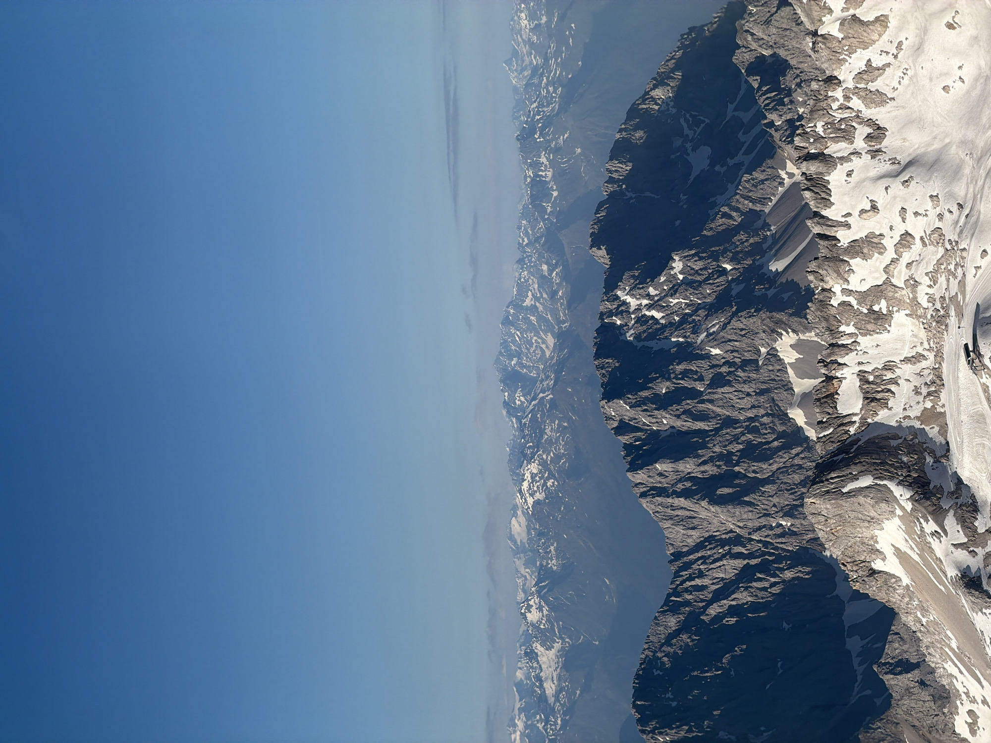 This is an aerial, wide-angle, eye-level shot of a mountain range, likely the Alps, under a clear blue sky. The foreground features a jagged, rocky mountain peak with patches of snow. Behind this, layers of more distant mountain ranges are visible, also covered in snow, receding into a hazy distance. The scene appears to be in a natural, mountainous environment, with no signs of a city. Given the context of Grainau, Germany, this is likely a view of the Zugspitze or a surrounding peak in the Wetterstein Mountains. There are no people or specific objects identifiable in the scene, only the natural topography of the mountains and the sky.