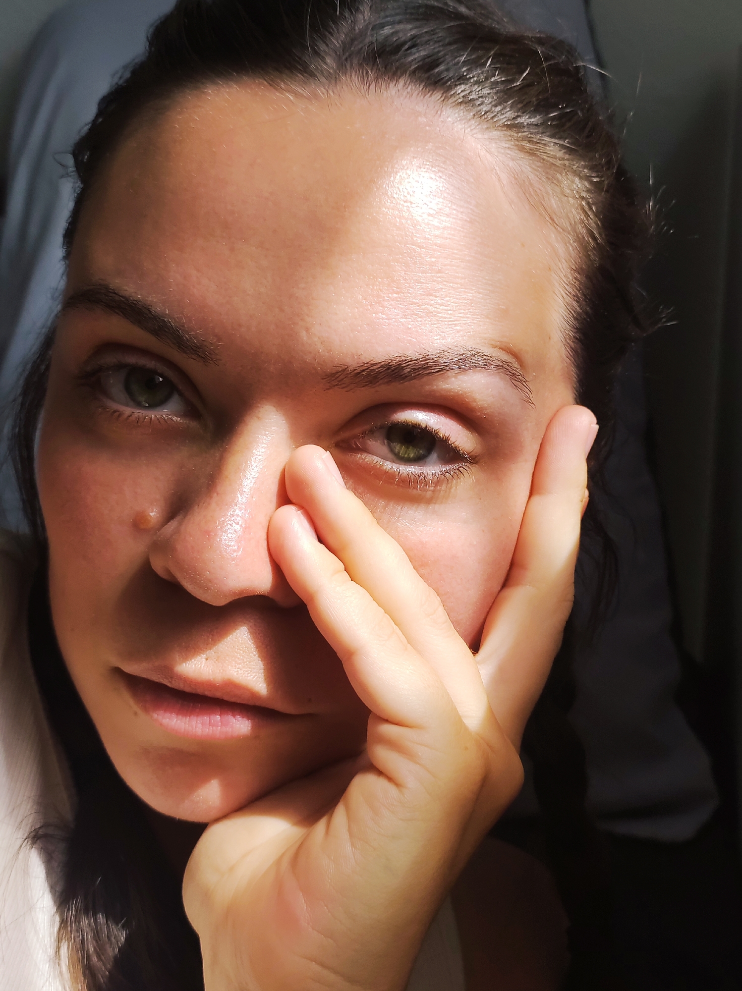 The image is a close-up, high-angle shot of a woman's face. Her left hand is placed across her cheek and mouth, obscuring part of her face. The woman has fair skin, green eyes, and dark hair pulled back. Sunlight illuminates the right side of her face, creating strong highlights and shadows. The background is dark and indistinct, with some lighter fabric on the left. The image appears to be taken indoors, possibly in a domestic setting. There is no visible information in the image that would allow for the identification of the city of Chaikovsky, Russia.