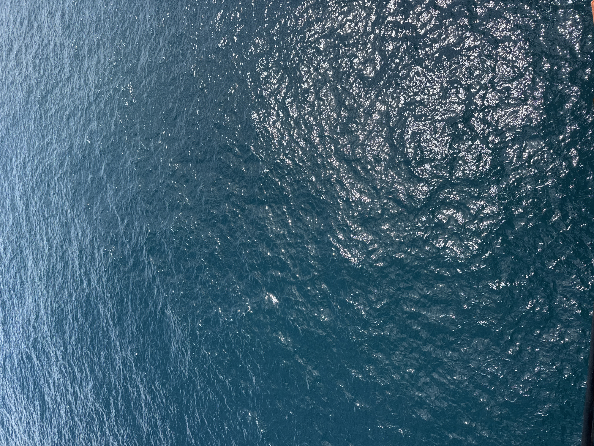 An overhead, medium shot captures the surface of the ocean. The water exhibits a deep blue hue, with ripples and waves creating a textured appearance. Sunlight glints off the water's surface in various spots, highlighting the movement and depth of the sea. The lower portion of the image shows a dark, horizontal railing, suggesting the vantage point is from a ship or elevated structure. The scene is devoid of any human presence or urban elements, portraying a natural seascape.