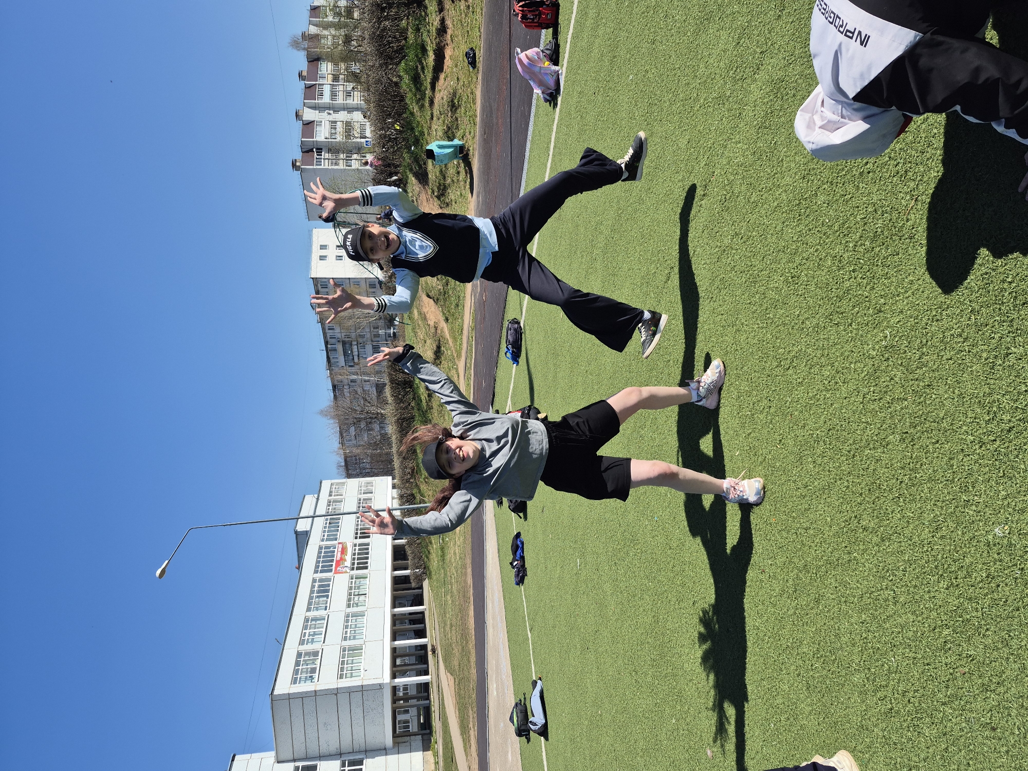 Two girls are jumping in the air on a green field in Chaikovsky, Russia. In the background are apartment buildings, trees, a running track, and other people. One girl wears a black cap, a blue and black vest, and black pants with sneakers. The second girl wears a gray jacket, black shorts, and sneakers. A street light is visible in the bottom left corner. A partial view of a person wearing a white jacket with black sleeves and a white cap is in the top right corner. There are various bags and items on the field.