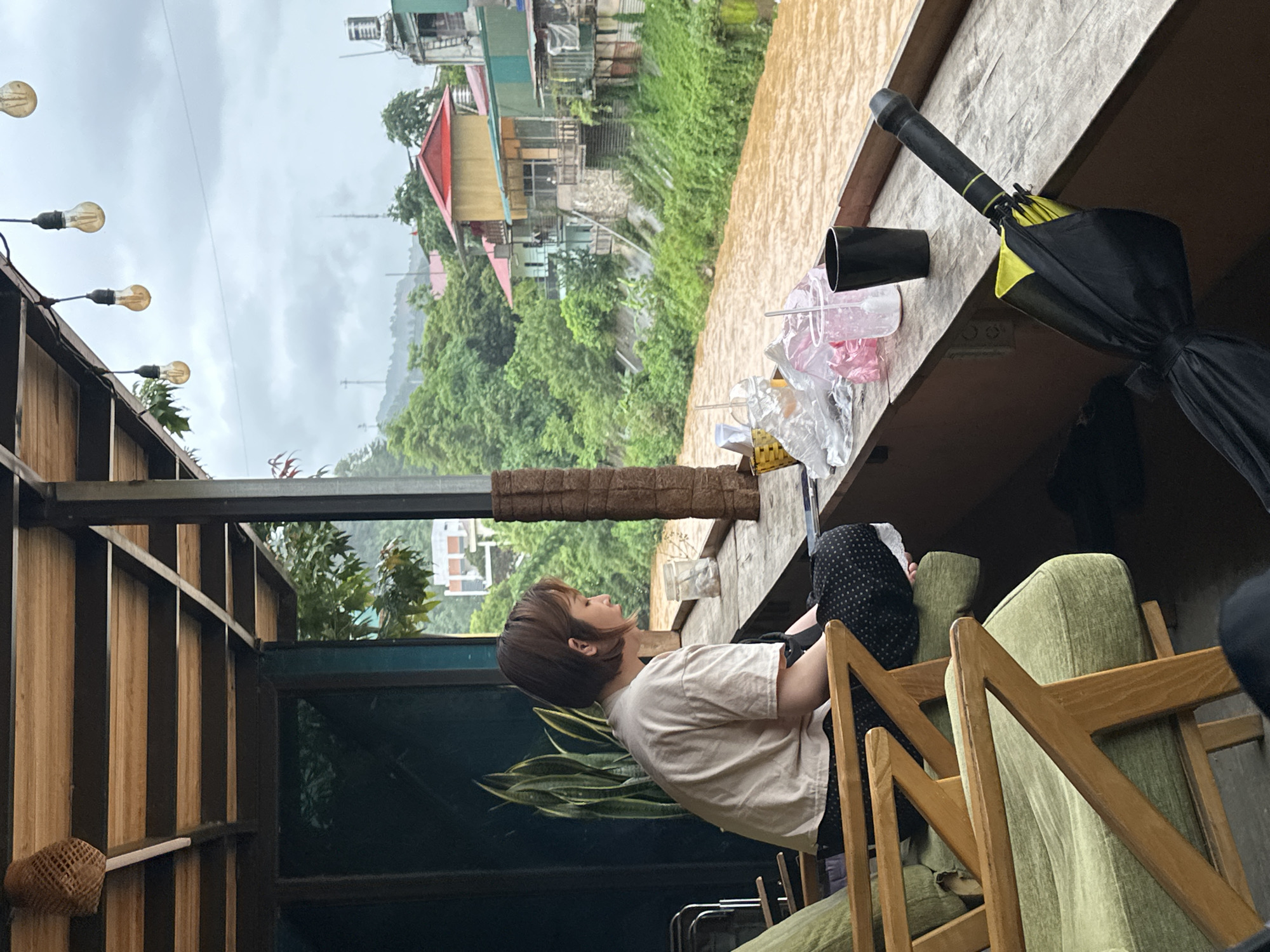 The image depicts a woman seated in a semi-outdoor setting, likely a cafe balcony or terrace, overlooking a picturesque mountainous landscape. The scene is set in what appears to be a cool, overcast day, possibly in Mù Cang Chải, Vietnam, given the lush green hills and type of architecture.
The main subject is a woman with short, light brown hair, wearing a plain light-colored t-shirt. She is seated on a light green cushioned chair, looking calmly towards the distant view. Part of another person, wearing a black and white patterned head covering, is partially visible behind her.
The immediate surroundings include wooden slatted walls and sturdy metal beams forming the structure of the seating area. String lights with exposed, warm-toned bulbs are strung along the overhead beams. On a nearby ledge to the right, a black, folded umbrella with yellow interior accents is stored, alongside some plastic containers and bags. A woven basket is attached to one of the wooden walls. A large snake plant adds a touch of greenery to the interior space.
Beyond the balcony, the background reveals a valley filled with dense green vegetation and trees, with several houses and buildings, some featuring reddish roofs, scattered throughout the hills. The sky is uniformly gray and cloudy, obscuring the mountaintops and contributing to the serene, slightly muted atmosphere. There is no visible text in the image.