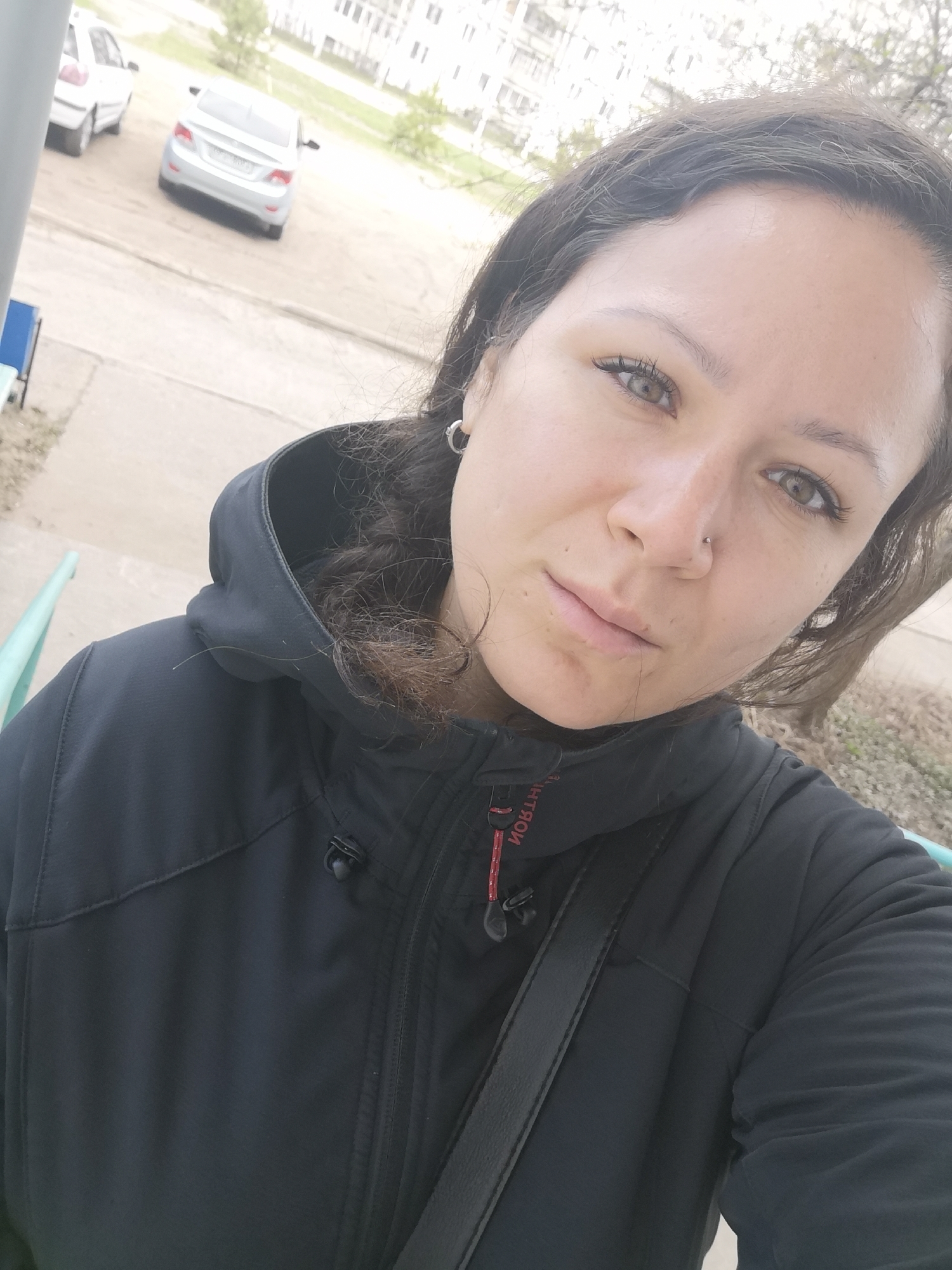 A selfie captures a woman outdoors in Chaikovsky, Russia. She has long, dark, wavy hair, and her light-colored eyes, possibly hazel or light brown, are focused directly on the camera. A small silver stud adorns her left nostril, and a silver hoop earring is visible on her left ear. She is dressed in a black hooded jacket, which appears to be made of a functional, possibly weather-resistant material. A black shoulder bag strap crosses her body. Her expression is neutral and composed.
The background reveals an outdoor urban or suburban setting with a light-colored ground, likely concrete or pavement, and some sparse vegetation. Two cars are parked in the distance: a white sedan partially visible on the left and a silver sedan in the center. A multi-story building is faintly discernible further back.
The lighting indicates daytime, likely overcast, as there are no strong shadows and the scene is uniformly bright. A red tag with reversed text, possibly "MONTON" or "MONTI", is visible on the zipper pull of her jacket.