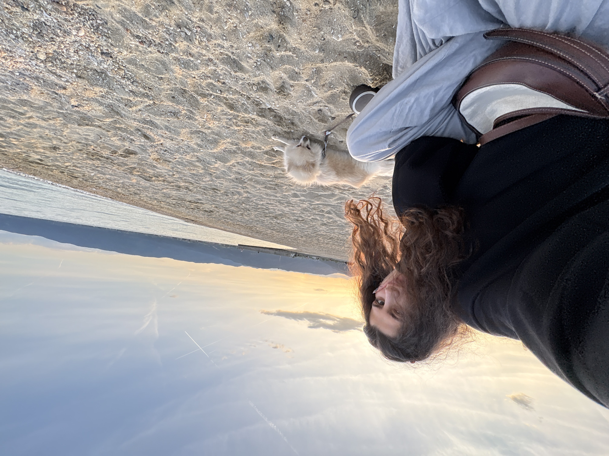 A person with long curly hair is taking a selfie at dusk on a beach in Σταυρός, Greece. The sky is visible with some clouds. A golden retriever dog is lying on the sandy beach near the person's legs and a beige bag. The sea and mountains are visible in the distance.
