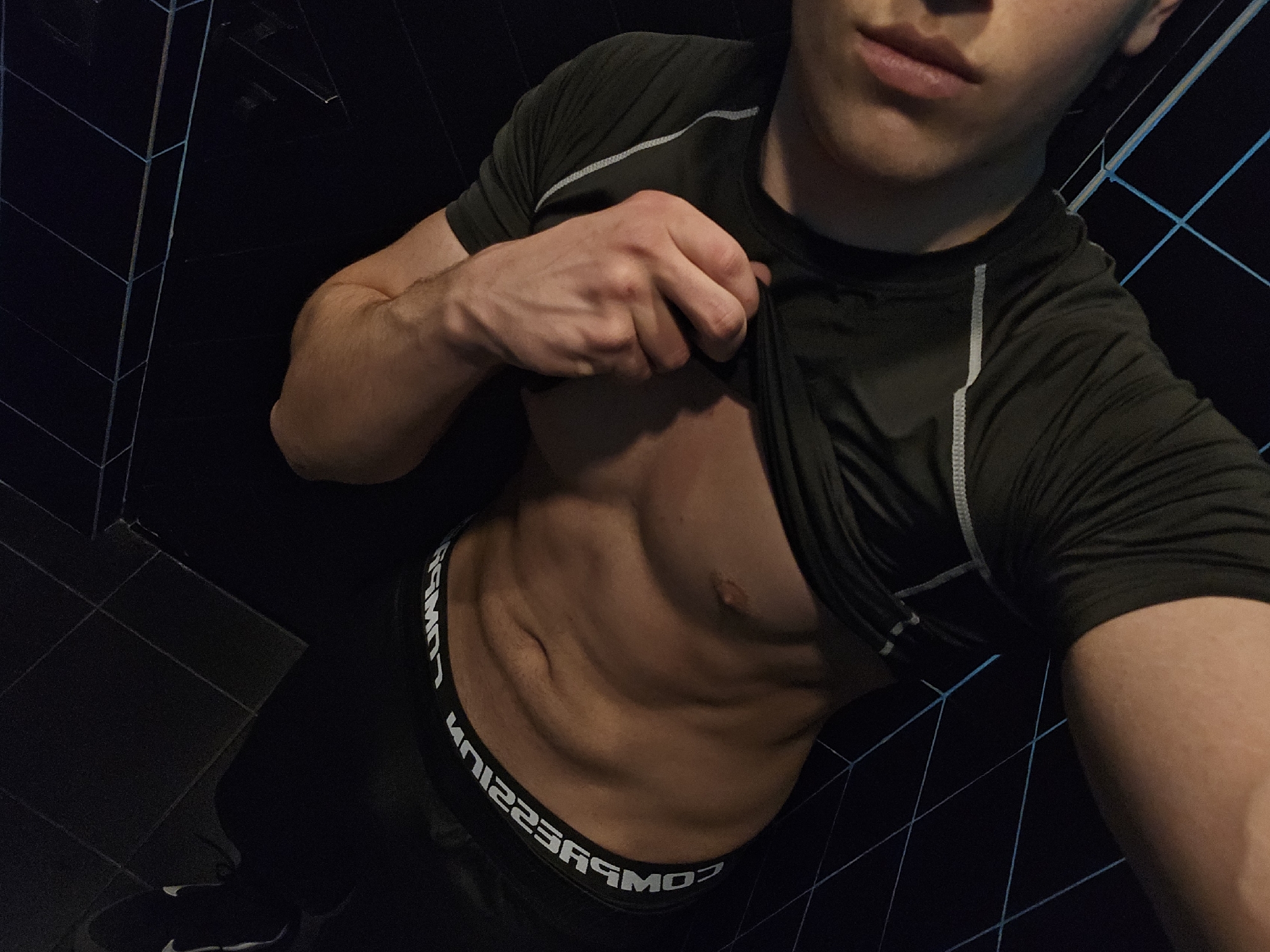 A medium shot depicts a person indoors, lifting the front of their dark-colored athletic shirt to reveal their abdomen. The person is wearing dark-colored athletic pants with "COMPRESSION" printed in white on the waistband. The background features dark tiles with light blue lines. The image does not provide information to determine the location in Yerevan, Armenia.