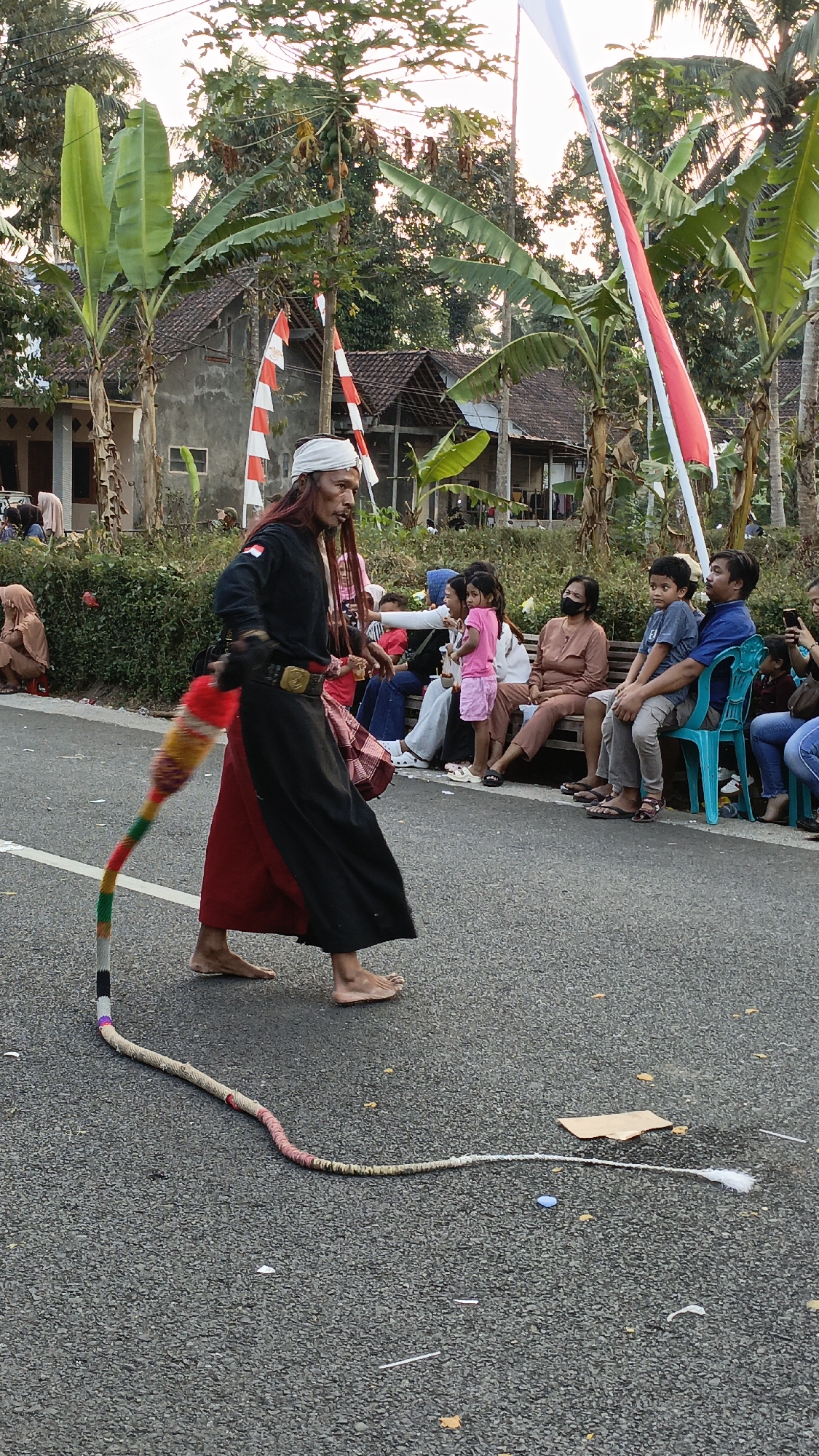In Krajan Lor, Indonesia, a man is seen performing outdoors, possibly during a festival or cultural event. He is wearing traditional attire, consisting of a black top with long sleeves, a red skirt or sarong, and a white headscarf. A long, colorful whip or rope, adorned with red, yellow, green, and white segments, trails behind him on the asphalt road. The man is barefoot and appears to be in motion, possibly mid-performance.
Behind the performer, a group of spectators are seated on a long bench and on chairs. They are diverse in age and gender. Some are wearing masks, indicating awareness of health precautions. In the background, houses with tiled roofs are visible, surrounded by lush green trees, including banana trees. Indonesian flags, red and white, are displayed on poles, further suggesting a celebratory or national event. The overall scene captures a moment of cultural expression and community gathering in a semi-rural Indonesian setting.
