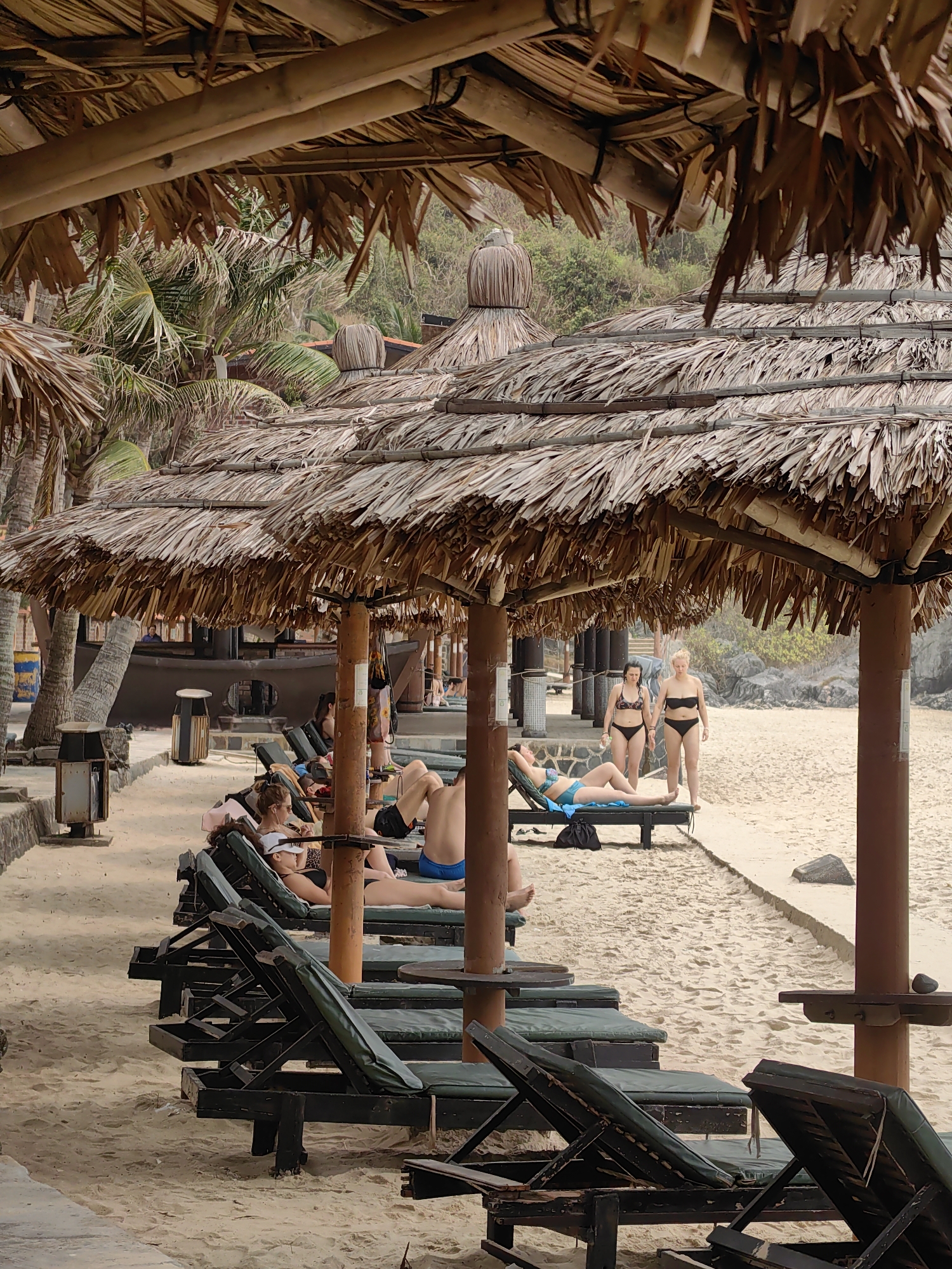 The image depicts a beach scene, likely a resort area, in Hải Phòng, Vietnam. In the foreground, a row of dark wooden lounge chairs with green cushions is set on the sandy beach. Several people are reclining on the chairs, some appearing to be sunbathing. A woman in a bikini is visible on a lounge chair further back, while a few others can be seen in swimwear near the edge of the sand. Two women are standing on the beach in bikinis. The beach is under the shade of thatch-roofed umbrellas supported by wooden poles. Palm trees and other foliage are visible in the background, along with structures such as a bar. The lighting suggests either an overcast day or that the sun is high in the sky, casting soft shadows.