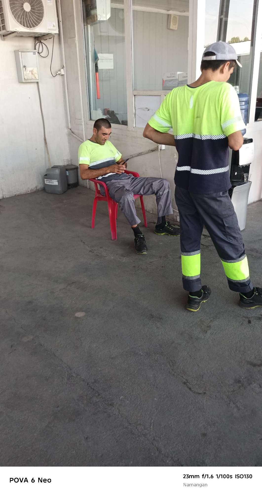 In Namangan City, Uzbekistan, a scene unfolds in what appears to be an outdoor service area or workshop. Two men are present. One man is seated on a red plastic chair, holding a mobile phone in his hands and looking down at it. He is wearing a light green t-shirt with horizontal navy and white stripes across the chest and sleeves, and grey trousers. The other man is standing, seen from the back, wearing a similar light green t-shirt with navy and white stripes, and dark grey trousers with bright lime green reflective bands around the lower legs. He also wears a grey baseball cap. Behind the seated man, there are grey bins. To the right of the standing man, a white water dispenser unit is visible. The environment is paved with concrete, showing some cracks and wear. An air conditioning unit is mounted on the wall in the background, along with windows and a doorway. The lighting suggests it is daytime.
