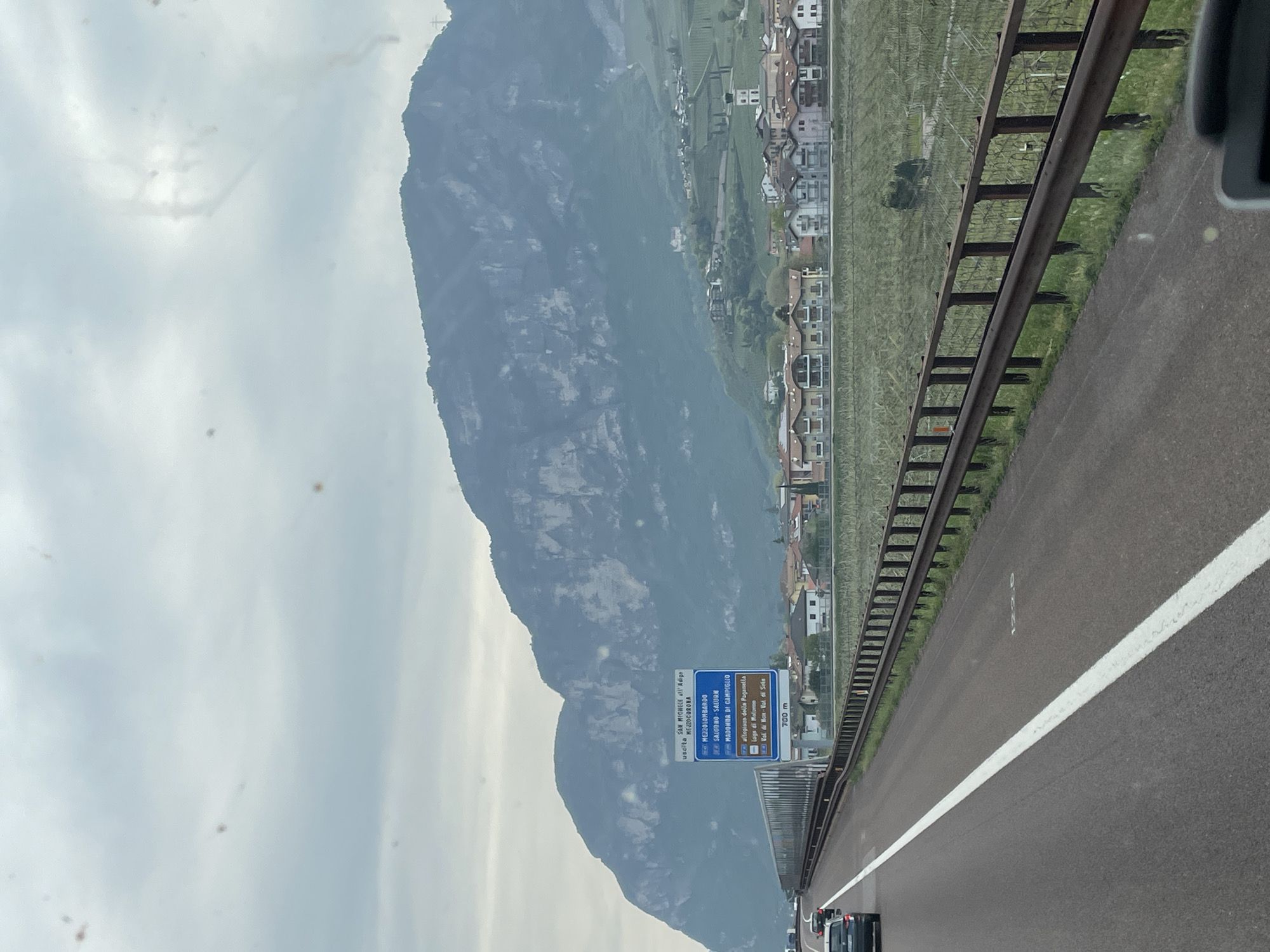 The image shows a view from a vehicle traveling on a highway near Nave San Rocco, Italy. In the background, a large mountain rises to the left of the frame. A town with closely packed buildings is situated between the mountain and the highway. A metal guardrail runs parallel to the highway. Road markings are visible on the asphalt. A blue and brown road sign indicates exits for various locations including San Michele all'Adige, Mezzocorona, Mezzolombardo, Salorno Salurn, Madonna di Campiglio, Altopiano della Paganella, Lago di Molveno, and Val di Non-Val di Sole, with a distance of 700 meters. Several cars are visible in the distance. The sky is overcast, and the overall lighting is subdued.