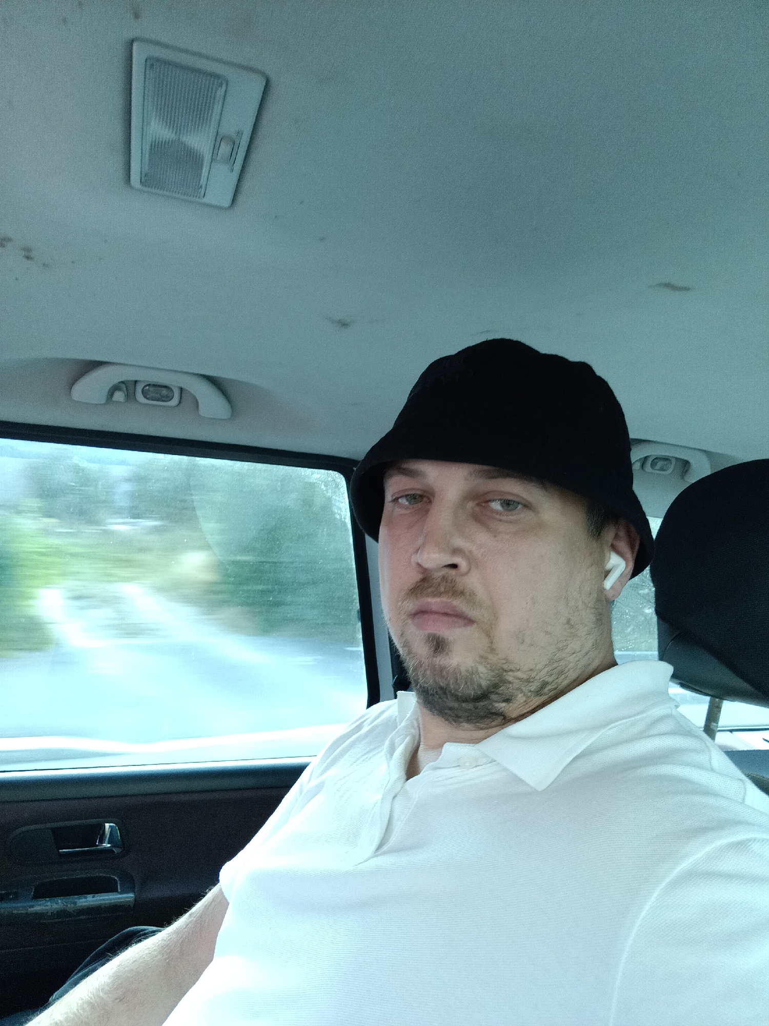 The media file is a selfie taken inside a vehicle, likely a car, showing a man with light skin, a beard, and wearing a black bucket hat and a white polo shirt. He has a neutral expression and is wearing white earbuds. The interior of the car is visible, including the ceiling with a light fixture and a grab handle, and the door panel with a window. Through the window, a blurry landscape of green trees and a road can be seen, indicating the vehicle is in motion. The lighting suggests it is daytime. The scene is consistent with being in Prague, Czechia, given the provided location context. There is no visible text in the image.