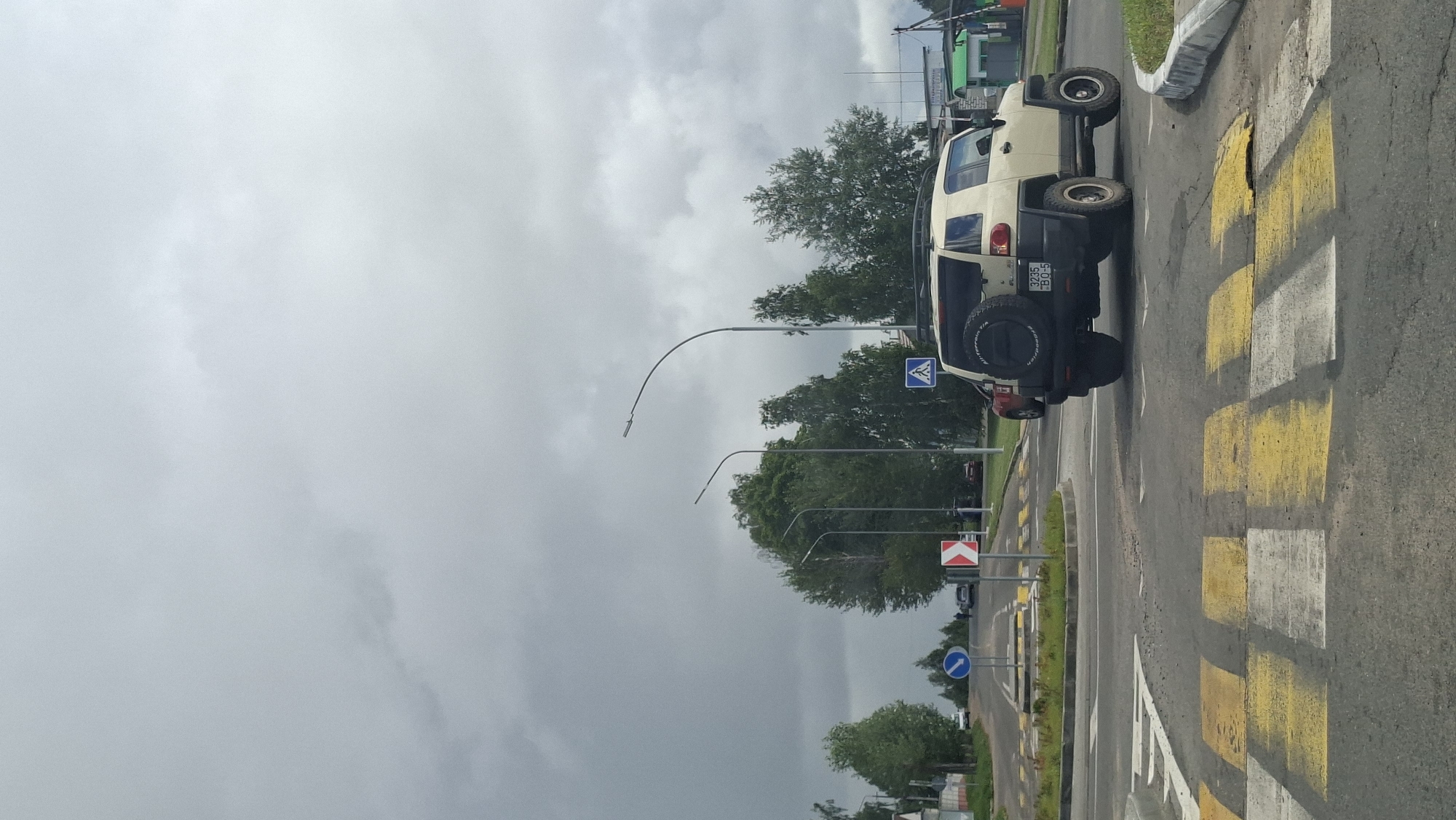 The image shows a beige SUV, possibly a Toyota FJ Cruiser, positioned on a roundabout at a crosswalk in Myadzyel, Belarus. The car is facing away from the viewer and appears to be in motion or preparing to move. The sky is overcast with heavy clouds, suggesting potential rain or stormy weather.
In the foreground, a pedestrian crossing is marked with alternating yellow and white stripes. The roundabout itself is circular with a grassy central island. Several traffic signs are visible, including a blue circular sign with a white arrow indicating a mandatory direction of travel, and a red and white chevron sign indicating a curve to the right.
Beyond the roundabout, trees and streetlights line the road. In the distance, other vehicles and buildings can be seen. The overall impression is of a typical intersection on a cloudy day in a suburban or semi-urban area. The environment appears to be a road intersection with pedestrian crossing, roundabout, trees, and streetlights. The weather is overcast.