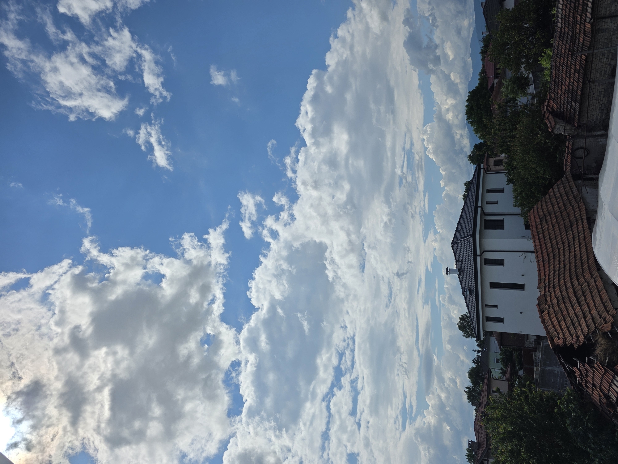 The image presents a daytime view in Yagodovo, Bulgaria, featuring a bright blue sky dotted with cumulus clouds. On the right side of the frame, a white building with a dark roof is visible, partially obscured by trees and rooftops of neighboring houses. Some trees, buildings, and roof tops fill the scene with greenery. Mountains appear in the far distance behind the buildings.