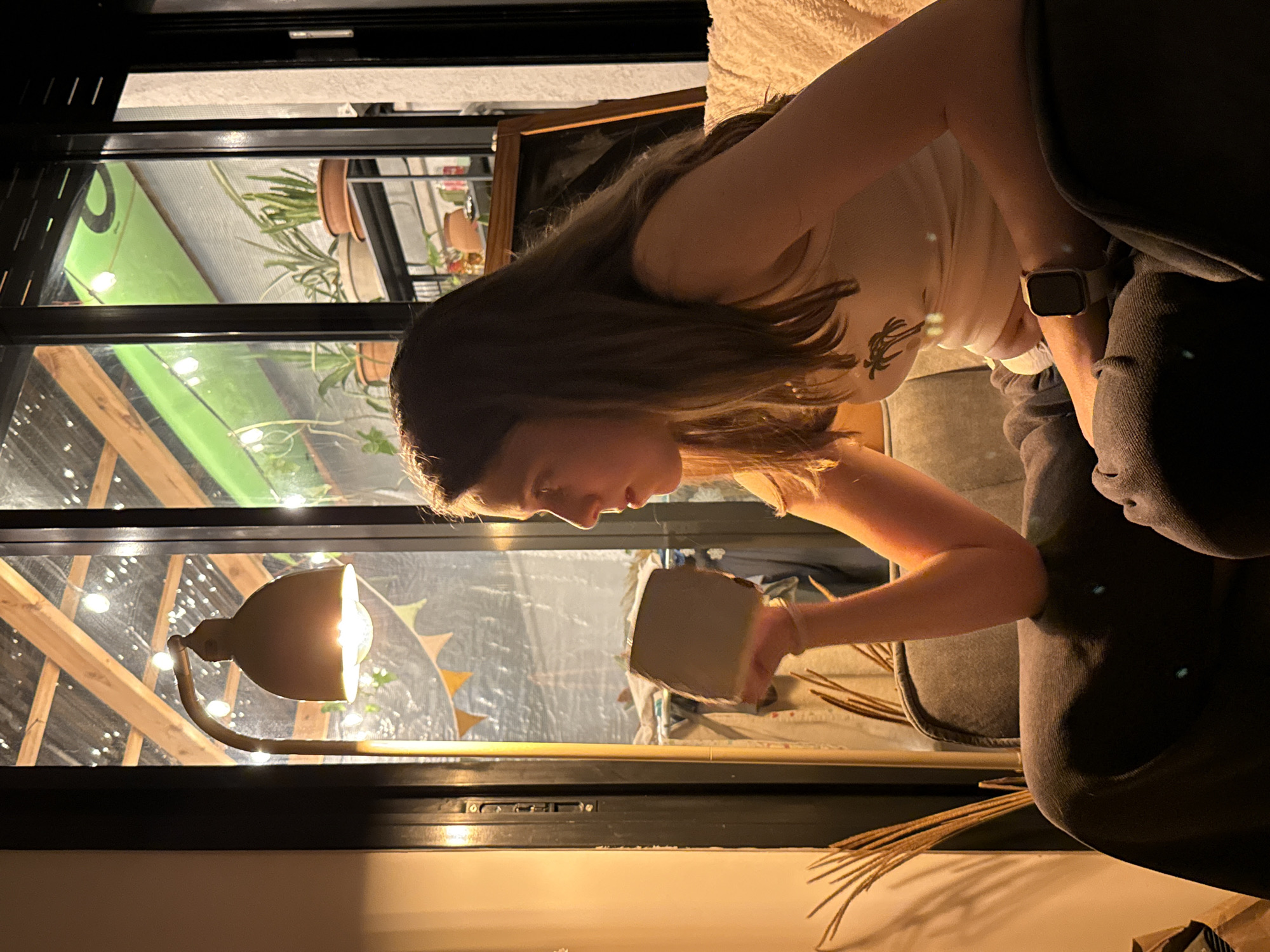 A young woman is depicted indoors, seated and viewed in profile, looking down as she holds a grey mug in her right hand. She has long brown hair and is wearing a light-colored tank top with a dark graphic print, possibly a palm tree. A light-colored smartwatch is visible on her left wrist. She appears to be sitting on a dark-colored couch or chair, with a part of its arm visible on the right.
The scene is set at night, with illumination provided by artificial light sources. A floor lamp with a beige/bronze metallic shade is prominent in the foreground, casting warm light. Through a large window or glass partition, an outdoor or semi-enclosed patio area is visible. This area contains several potted plants, including succulents and other green foliage. Wooden beams form part of a roof structure above, and numerous string lights are strung across the space, creating a festive atmosphere. Colorful triangular pennants/bunting are also visible as decorations. In the upper left background, through the glass, a bright green sign with a partial black, stylized letter, possibly an 'O', can be seen. Dried reeds or plant stalks are present on a surface in the lower right foreground. The overall ambiance is cozy and relaxed, suggesting a quiet evening in Tel-Aviv, Israel.
