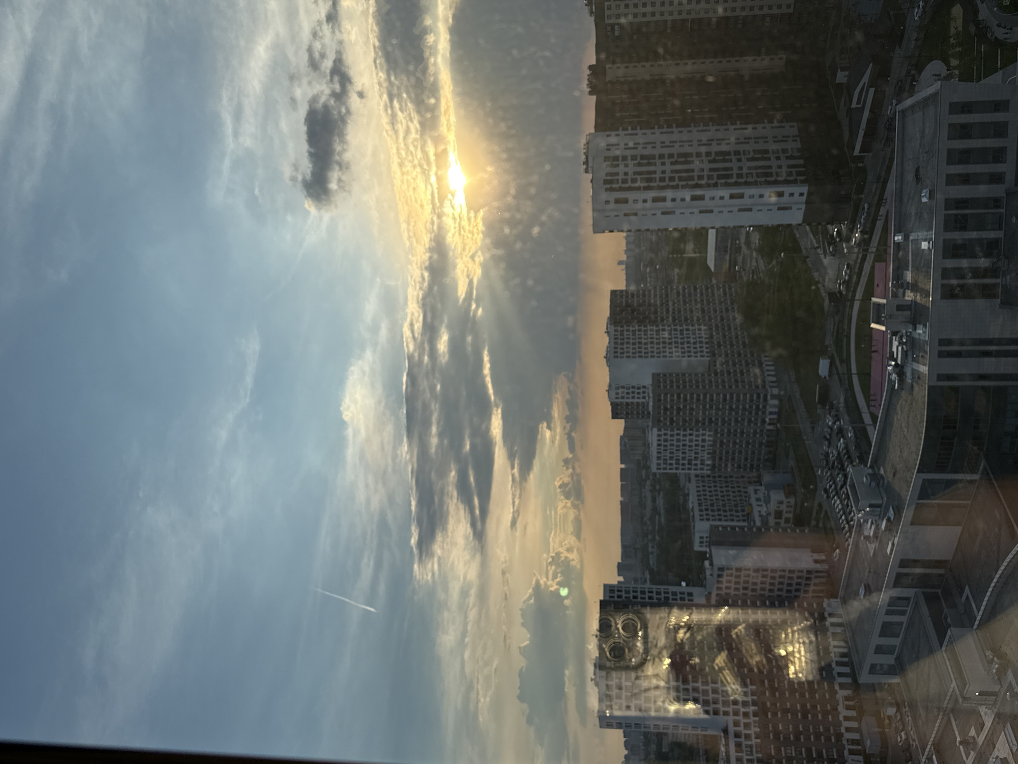 The image depicts an urban landscape at sunset, observed from a high vantage point, likely a tall building in Lyubertsy, Russia. The view includes residential buildings and the sky. The sky is filled with clouds, illuminated by the sun setting, creating streaks of light. The residential buildings exhibit a repetitive pattern of windows and architectural elements. The reflection of the city and sky is visible on a window pane with a cell phone on the window. The city seems to have a modern layout.