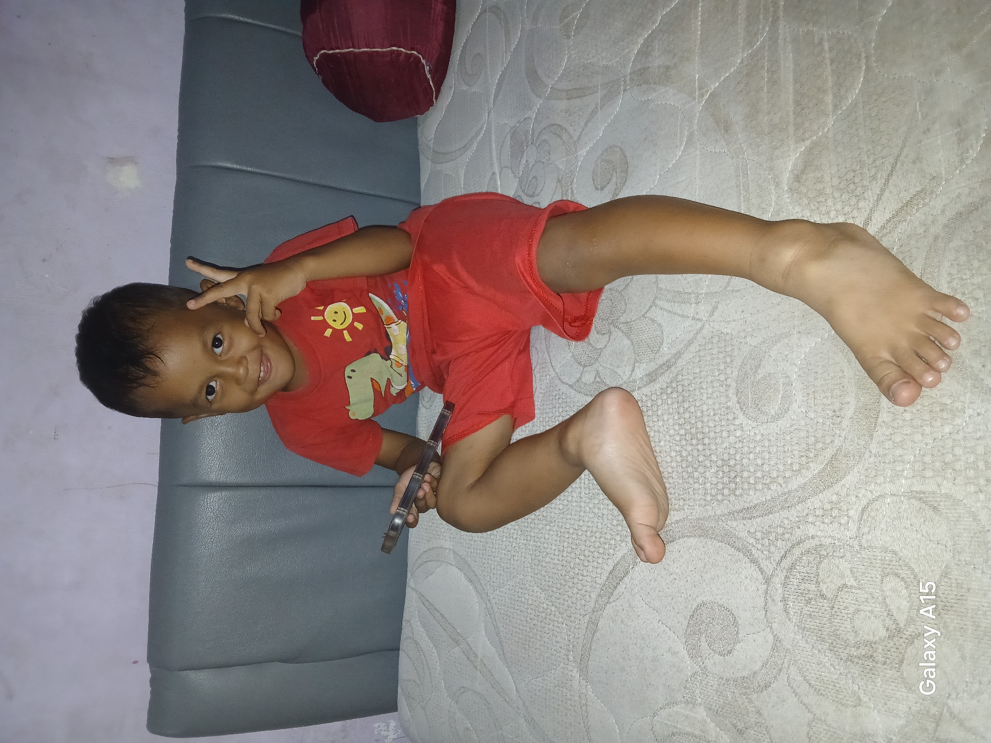 In Bekasi, Indonesia, a young boy is seated on a mattress. He is wearing a red t-shirt with a graphic of a dinosaur and a smiling sun, and red shorts. The boy is holding a silver object, possibly a phone, in his left hand and has his right hand raised in a peace sign. He is looking at the camera and smiling. The background consists of a gray headboard and a light purple wall. A dark red pillow is visible on the right side of the frame. The mattress has a textured pattern. The bottom left corner of the image shows the text "Galaxy A15".