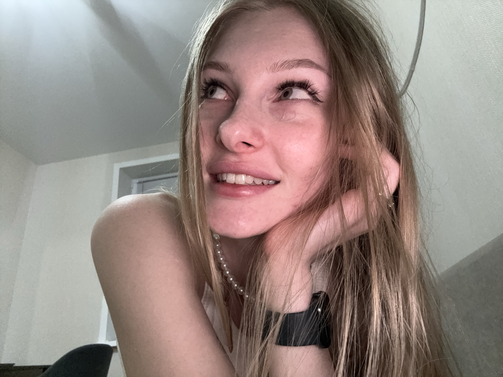 The image is a close-up, low-angle shot of a young Caucasian woman with long, light brown hair and fair skin. She is looking upwards and to her left, with her mouth slightly open, revealing her teeth. Her eyes are wide and light-colored, and she is wearing long, dark false eyelashes and subtle eye makeup. Her right hand is resting near her face, with her fingers gently touching her hair. She is wearing a white top and a pearl necklace. On her left wrist, she has a black watch. The background is a neutral-colored wall, with a hint of a window frame and a white ceiling. The lighting appears to be artificial, creating soft shadows. The image does not provide any discernible clues about the specific location within the city of Perm, Russia, or any specific urban context.