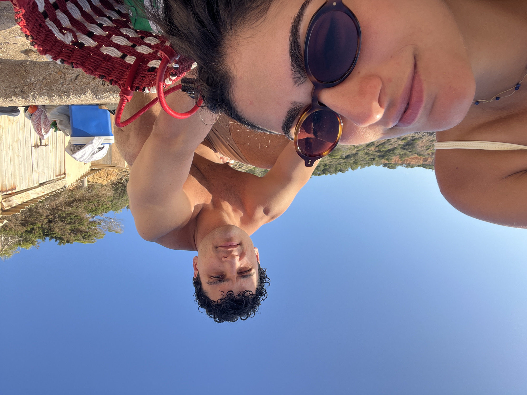This is a selfie taken outdoors on a beach, likely in Sant Josep de sa Talaia, Spain, given the clear blue sky and Mediterranean vegetation. In the foreground, a person is lying down, with their face close to the camera and slightly out of focus. They are wearing sunglasses and a white bikini top, with a necklace visible around their neck. Behind them, a shirtless man is kneeling, looking towards the camera. He has curly dark hair. In the background, there's a rustic building with wooden doors, a blue cooler, and some bags. The ground appears to be sand and stone. The overall impression is a casual, sunny day at the beach.