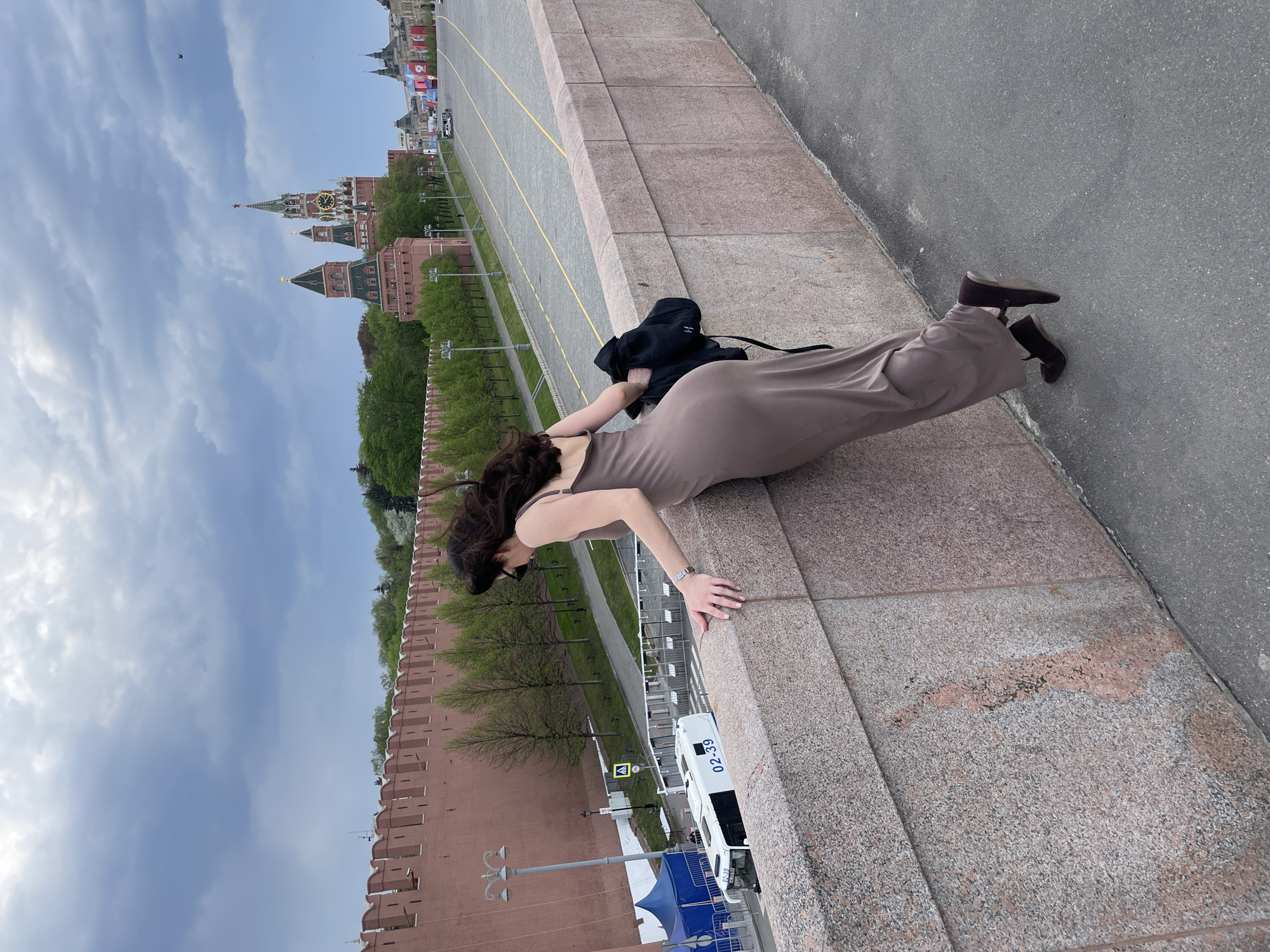 A woman in a long dress is leaning over the edge of a stone wall in Moscow, Russia. She is wearing brown shoes and has a black backpack beside her. The Kremlin and Red Square are in the background. A police van is parked below, next to a blue tent.
