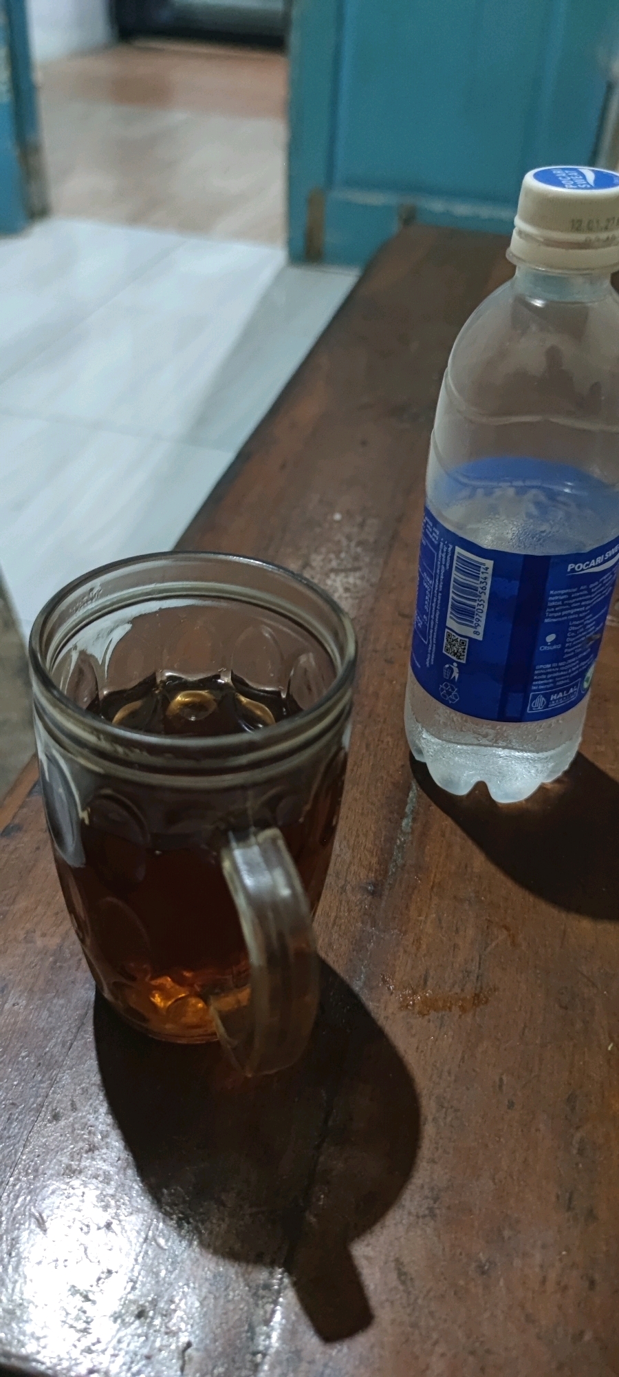 In Gambarsari, Indonesia, a glass mug filled with a dark liquid, likely tea, sits on a wooden table. Beside it, a partially consumed bottle of Pocari Sweat is visible, its label indicating it is a sports drink. The lighting suggests it is daytime, possibly indoors, with a plain background. The scene is static, with no human presence or activity depicted.