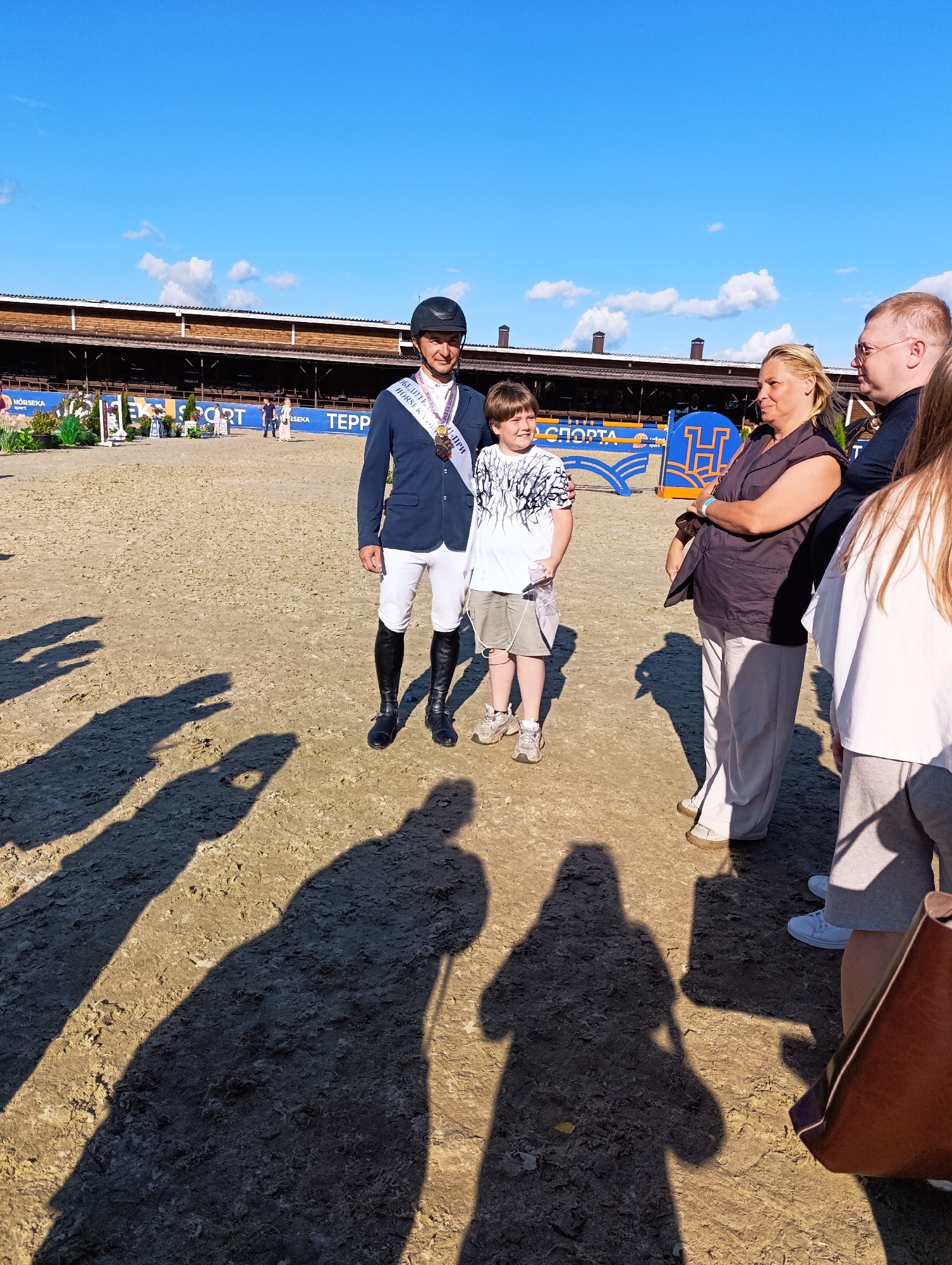 The image depicts an outdoor event at a horse riding arena, likely in Ivanovskoe, Russia, given the context. In the foreground, a group of people are standing on a dirt surface. A man in a dark blue blazer, white riding pants, black boots, and a helmet, wearing a sash and medal, is standing slightly off-center. To his left, a young boy in a white t-shirt with a black graphic print and shorts is holding a bottle. To the right of the man, a woman with blonde hair wearing a dark sleeveless top and light-colored pants stands with her arms crossed. Behind her, a man with glasses and a dark shirt is partially visible. Further to the right, a young woman in a white top and grey pants is standing. Long shadows are cast across the ground from the people in the foreground, indicating a sunny day. In the background, a wooden structure resembling a grandstand or stable building is visible, with signage that includes "SPORT" and "TEPP". The sky is clear blue with scattered white clouds.