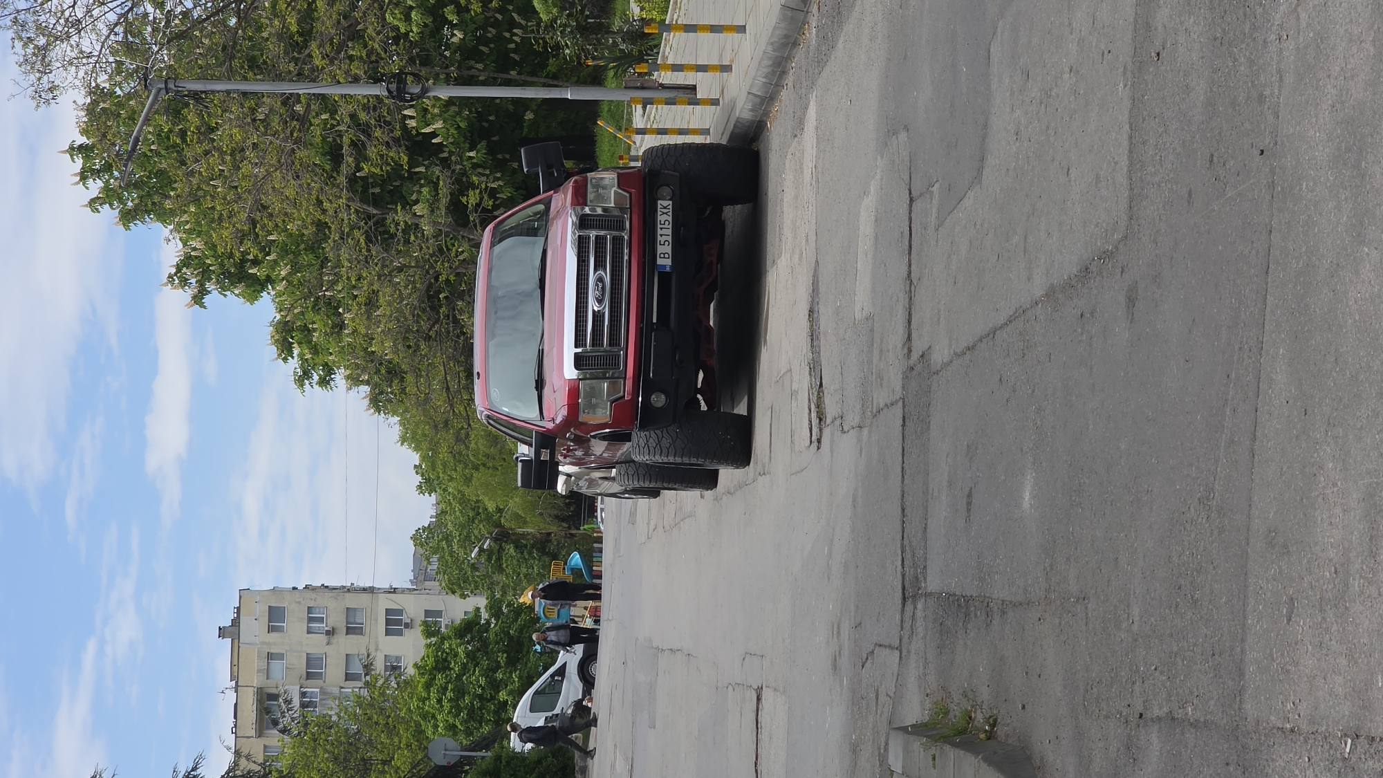 A red Ford pickup truck with a Bulgarian license plate reading "B 5115 XK" is parked on the side of a concrete city street. The scene shows an outdoor urban environment, with a multistory building and trees in the background, suggesting a residential area. Other vehicles and people are present in the distance. The location is in Varna, Bulgaria.