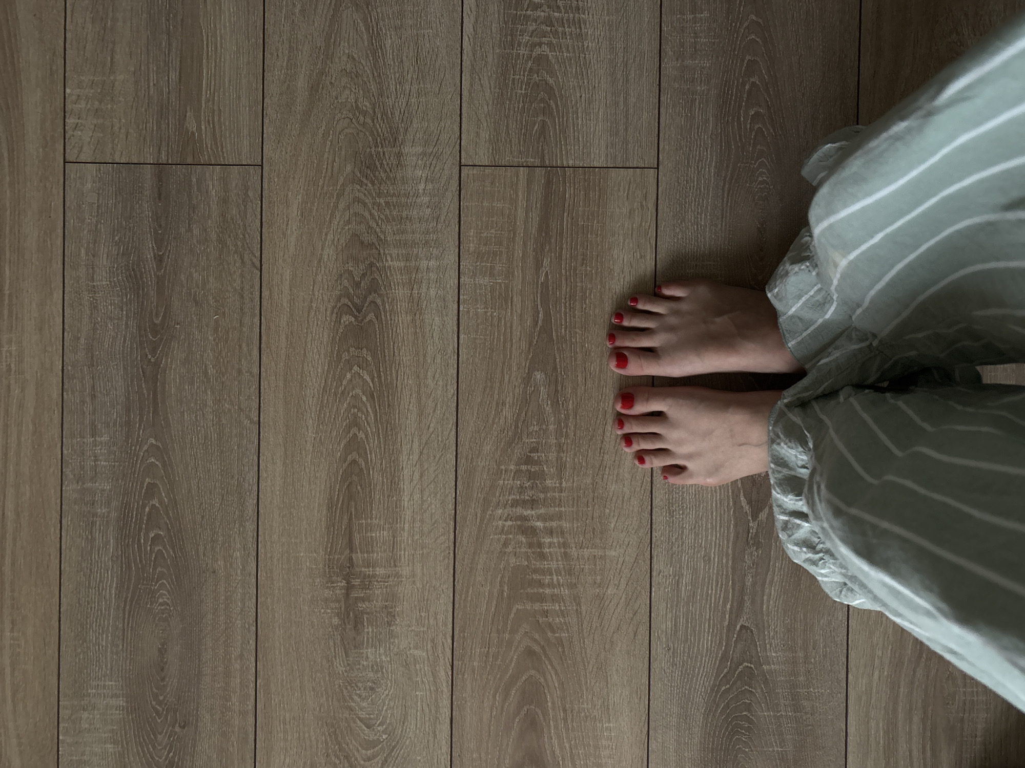 A top-down view of a person's feet and lower legs standing on a wooden floor. The feet are bare, with toenails painted red. The person is wearing light green pajama pants with white vertical stripes. The floor is made of wood-look laminate planks. The image was taken indoors, likely in a residential setting. There is no discernible location in the city of Zheleznogorsk, Russia.