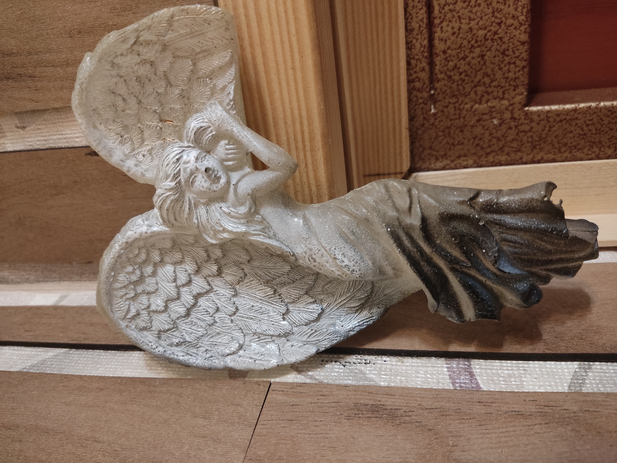 A white plaster angel figurine is affixed to a wooden doorframe in a residential interior. The angel has feathered wings, a flowing gown, and a delicate facial expression. The background consists of wooden paneling and what appears to be a textured door or wall surface. The image does not contain any information that would allow for specific location identification within Noginsk, Russia.