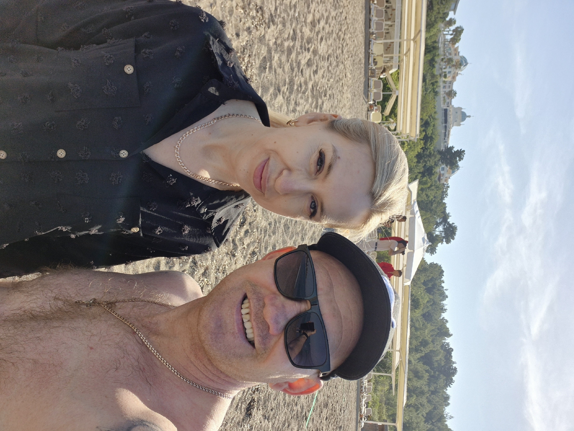 A selfie captures a couple on the beach in Alanya, Turkey. The woman, fair-skinned with blonde hair pulled back, wears a black blouse with light-colored buttons and a gold chain necklace. The man, also fair-skinned, is shirtless and wears sunglasses and a dark-colored cap. He also has a gold chain necklace. The beach sand is visible in the foreground and background. In the distance, there are beach umbrellas and lounge chairs and an elevated view of a building behind trees. The sky is blue with faint clouds.