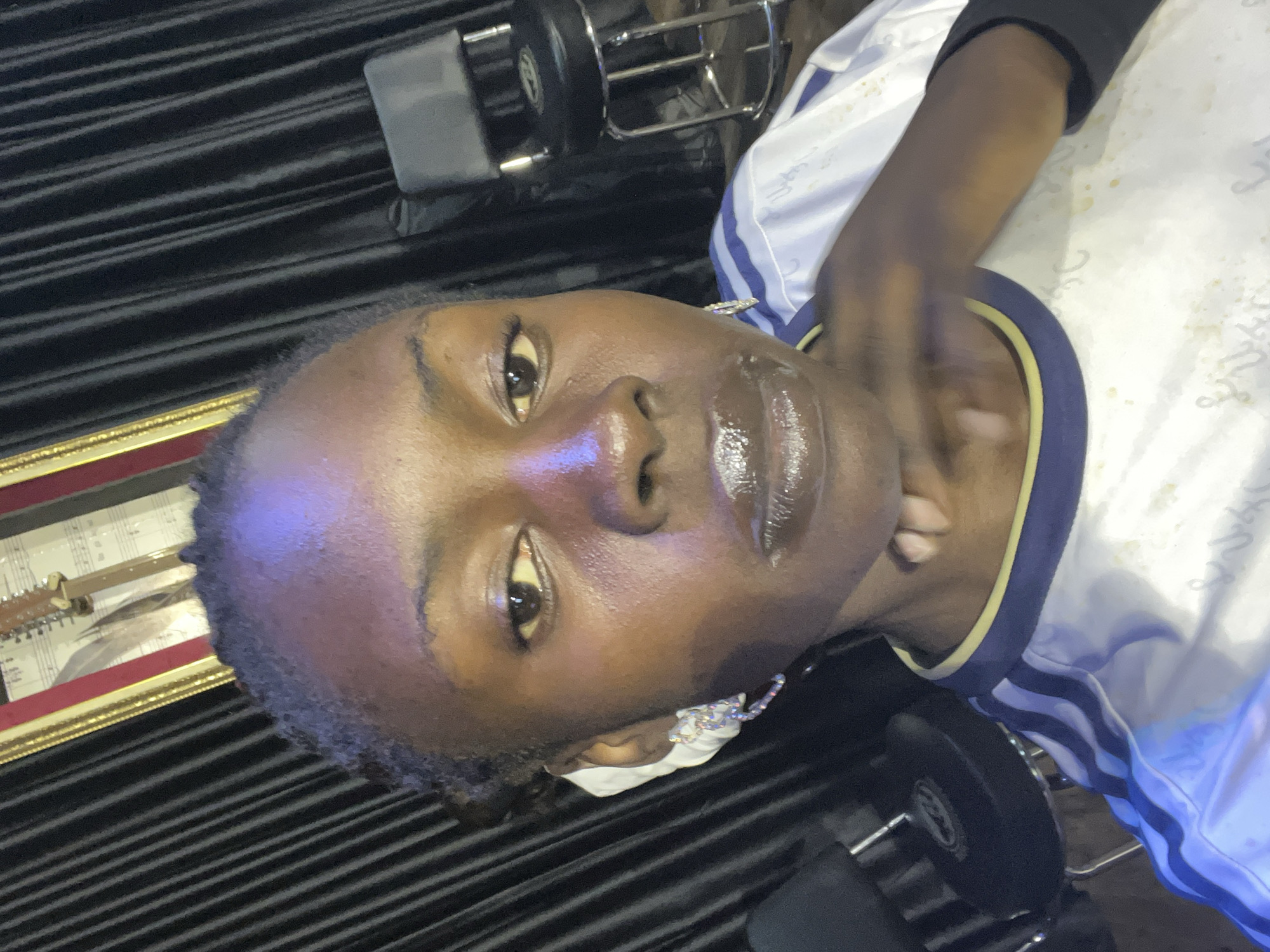 A young Black woman is captured in a close-up selfie, taken from a slightly low angle. She has dark skin, braided hair pulled back, and is wearing striking, sparkly earrings. Her lips are coated in a glossy, shimmering lip gloss, and she appears to be wearing makeup. Her right hand is raised to her chin, with her fingers gently resting on her jawline. She is wearing a white shirt with blue and yellow trim, and blue stripes on the sleeves. In the background, dark, pleated curtains are visible, along with framed artwork displaying musical notes and a guitar. To the right, a black stool with a metal frame is partially visible. The lighting in the scene is dramatic, with a noticeable purple and blue hue illuminating the subject's forehead and face. The overall setting suggests an indoor space, possibly a studio or a performance venue, and the aesthetic implies an artistic or creative environment.