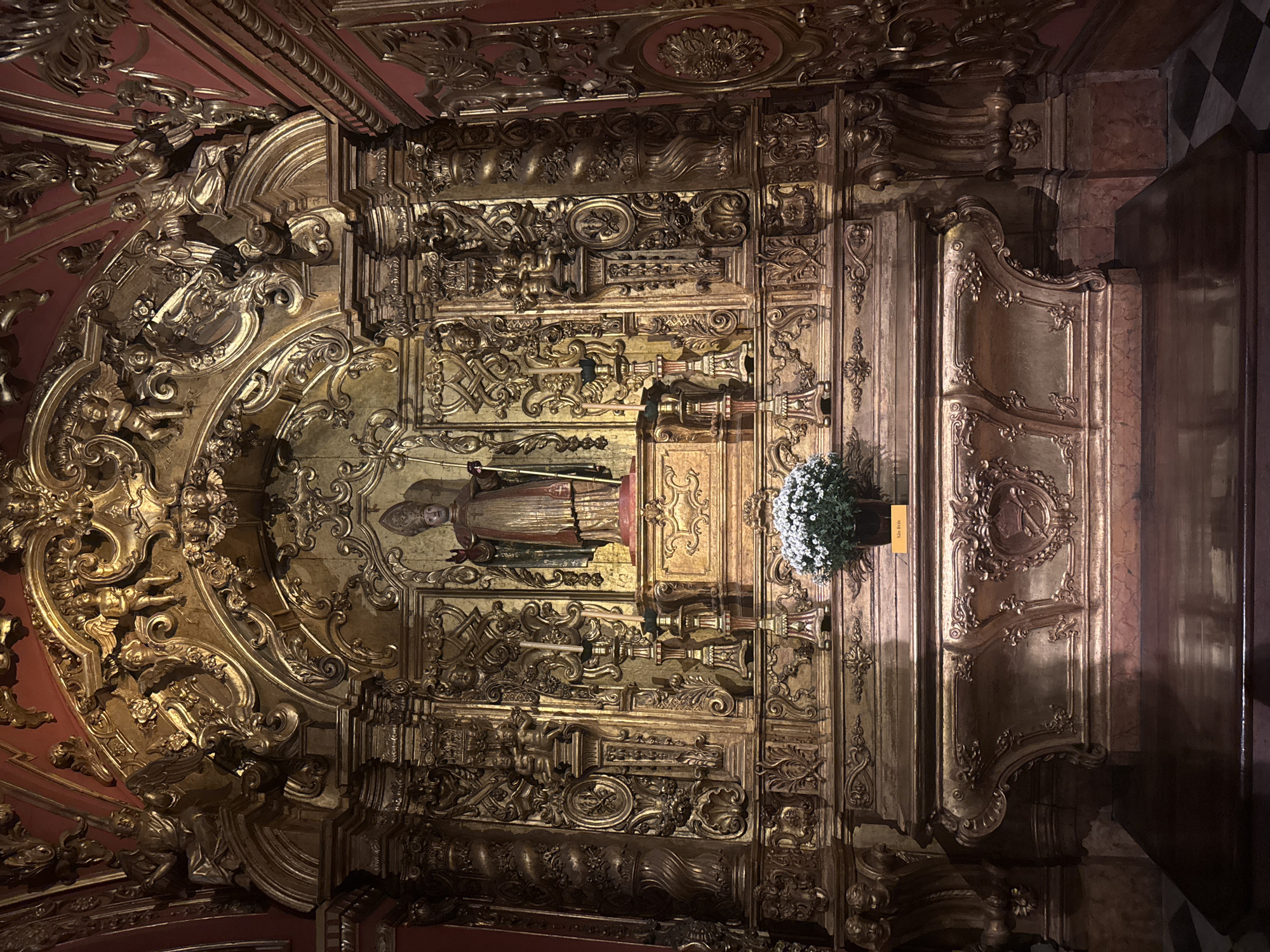The image shows a richly decorated altar within the Church of Saint Sebastian of the Capuchins (Igreja de São Sebastião dos Capuchinhos) in Rio de Janeiro, Brazil. The altar is an elaborate gilded wood retable featuring intricate carvings of cherubs, foliage, and architectural elements. At the center, above the altar table, stands a statue of a robed saint with a miter and staff. The altar table itself is also ornately carved and gilded, with decorative reliefs. A small bouquet of white flowers in a dark pot sits on the altar table, accompanied by a small yellow sign. The background walls are painted a deep red, contrasting with the golden ornamentation. The lighting is warm and somewhat dim, highlighting the textures and details of the gilded wood.