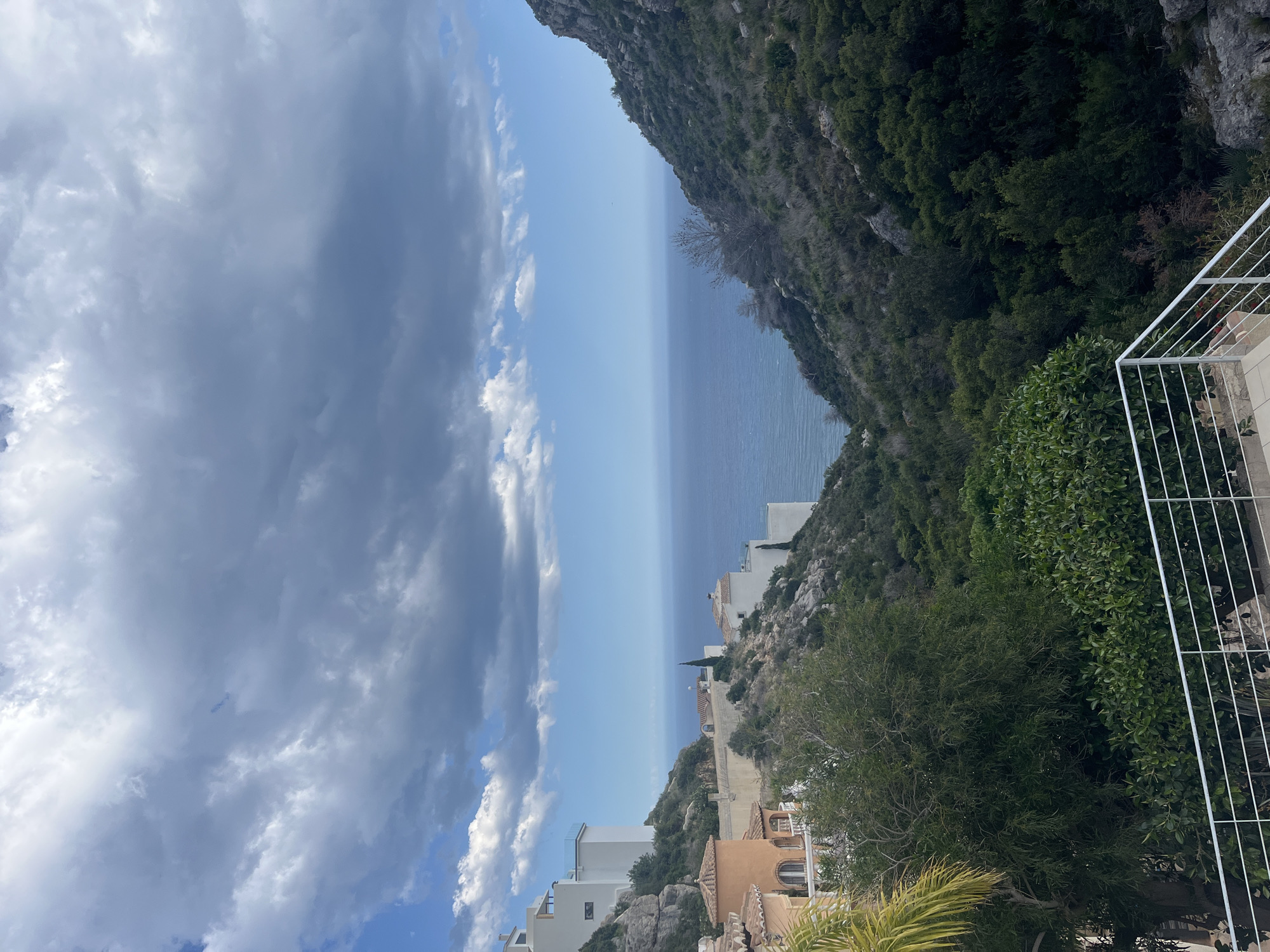 The image shows a view from a high vantage point, likely a balcony or terrace, in the coastal area of El Poble Nou de Benitatxell/Benitachell, Spain. The scene includes a rugged, green cliff face on the right side of the frame, dropping down to the Mediterranean Sea. Residential buildings, mainly white and light-colored villas, are nestled along the cliffside and further down the slope. A white railing is visible in the foreground, indicating the viewing point. The sky occupies a significant portion of the image, with a mix of dark, heavy clouds and patches of clear blue. The sea stretches across the horizon, transitioning from a lighter blue near the sky to a darker shade further out. The overall impression is of a coastal landscape with a combination of natural elements (cliff, sea, sky) and human development (buildings).