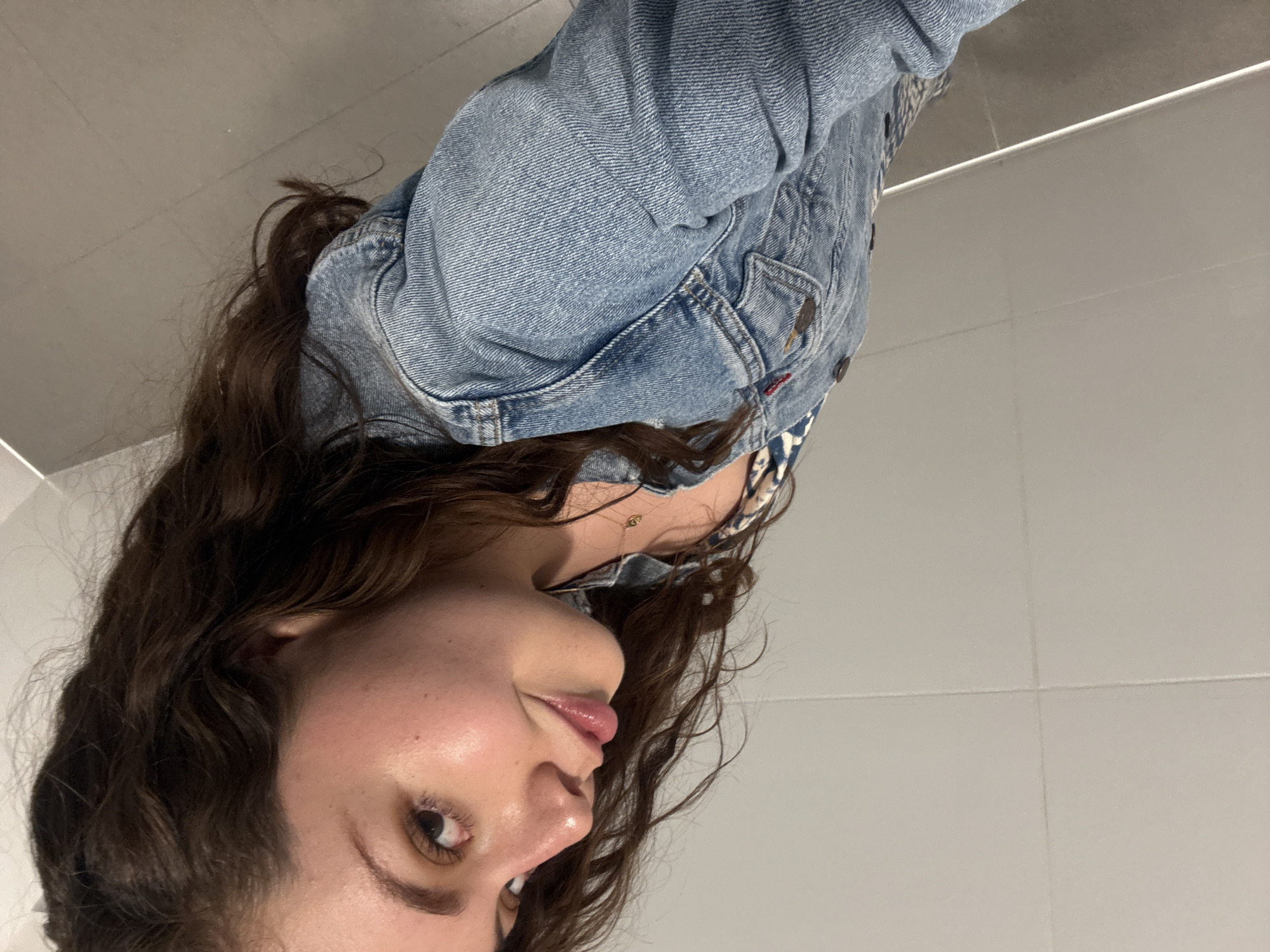 A young woman with brown curly hair and light skin is taking a selfie in what appears to be a public restroom. She is wearing a blue denim jacket over a patterned top. The background consists of white tiled walls. The location appears to be Mexico City, Mexico.