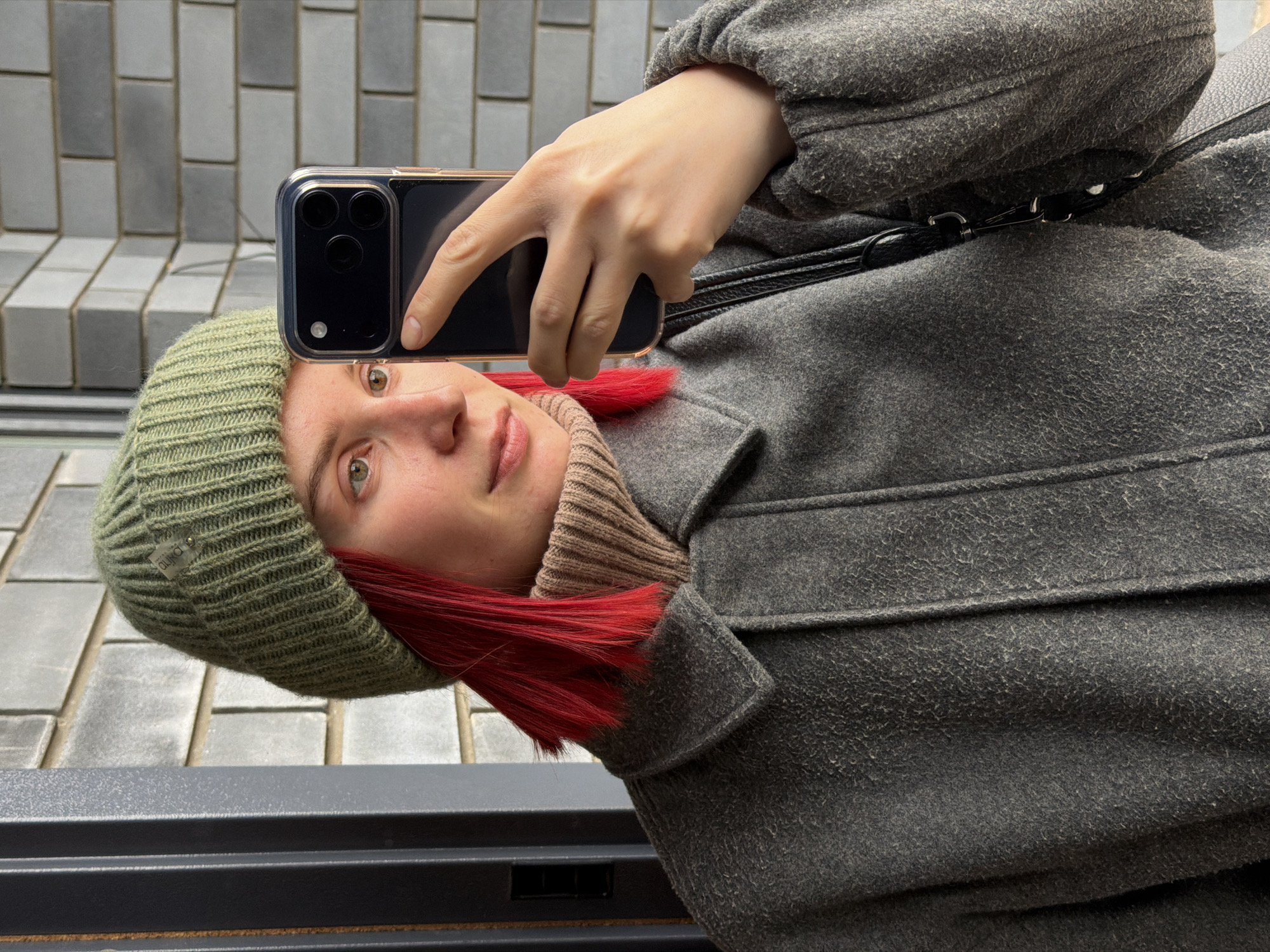 A young woman is taking a selfie with her smartphone. She is wearing a green knit hat, a beige turtleneck sweater, and a grey wool coat. She has dyed red hair. The phone case is clear. In the background are gray brick-like pavers. The photograph was taken in Chaikovsky, Russia.