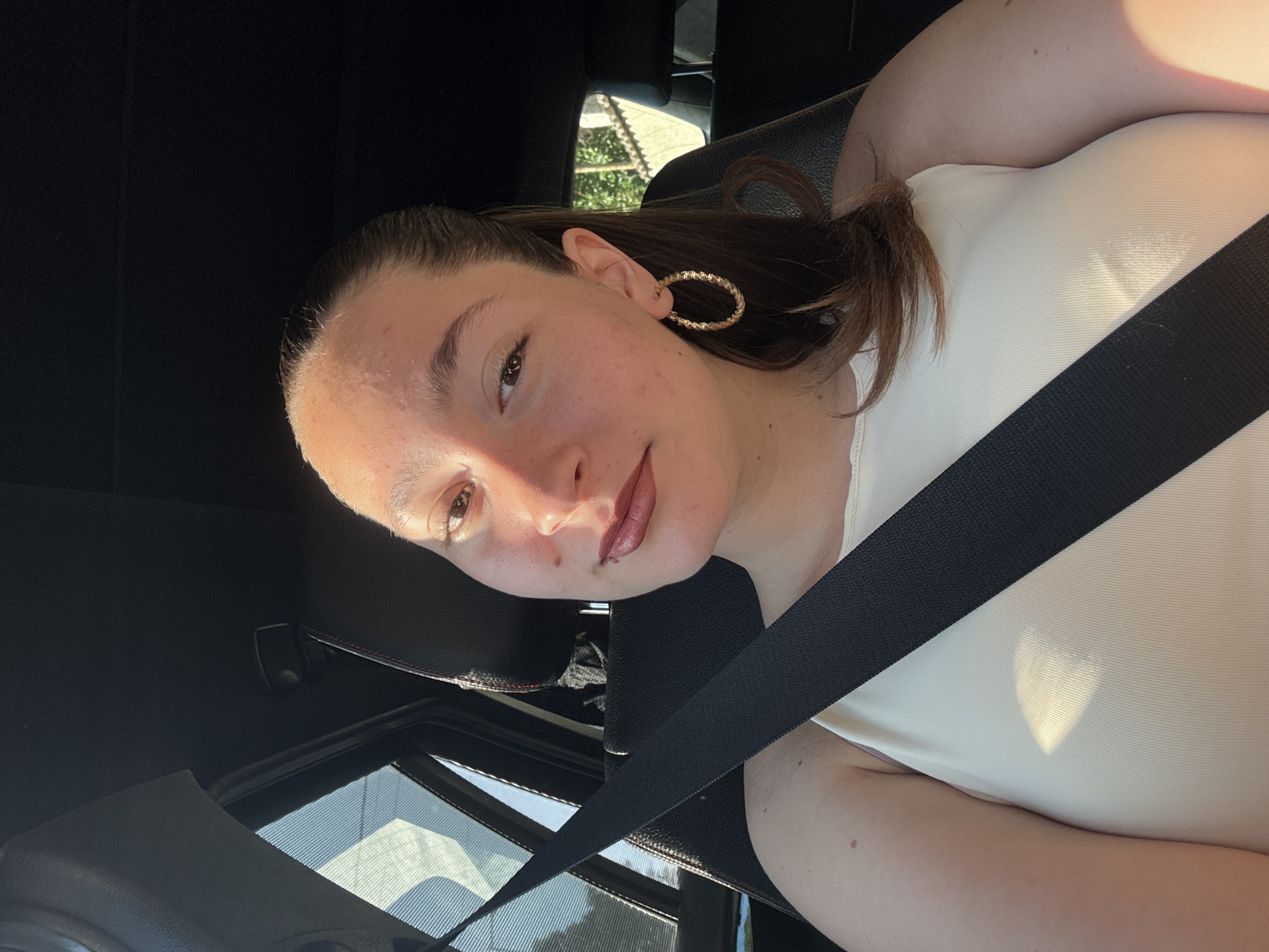 A young woman is sitting in the passenger seat of a car, likely in Sofia, Bulgaria, as indicated by the style of the car's interior and the surrounding foliage. She is wearing a white sleeveless top and large hoop earrings. A black seatbelt is diagonally across her chest. Sunlight casts a rectangular shadow on her top, and also highlights her face. She has brown hair pulled back into a ponytail and is looking at the camera. The car interior is dark, with a window on the left showing a blurred view of trees and buildings.