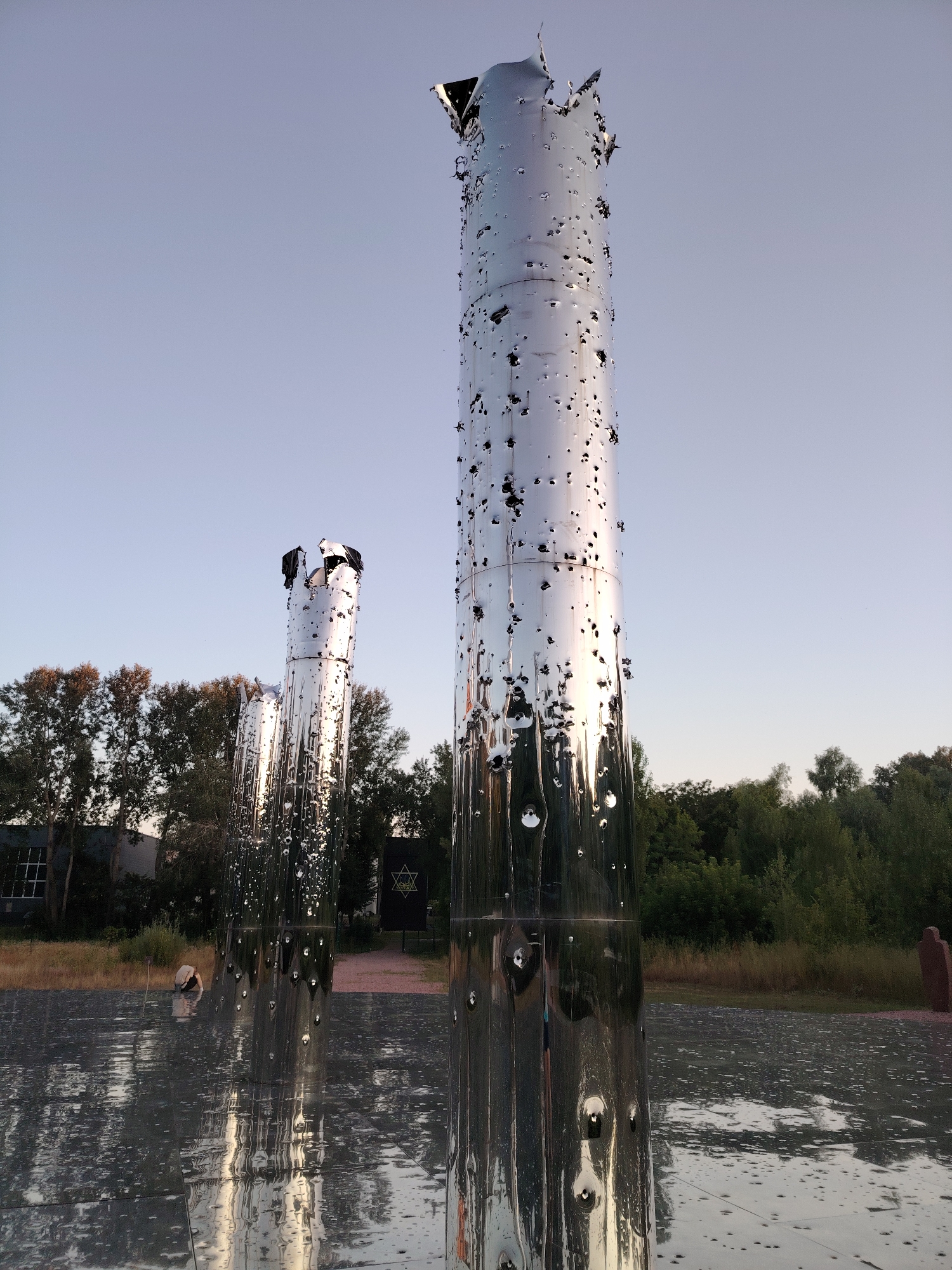This image depicts the "Columns of Memory" installation at the Babyn Yar Holocaust Memorial Center in Kyiv, Ukraine. The installation features two tall, cylindrical, mirrored structures that appear to be damaged or broken at the top, with numerous dark, irregular holes scattered across their reflective surfaces. The polished metal reflects the surrounding environment, including a clear sky, trees, and a building with a Star of David emblem. The structures are situated in a reflective plaza, likely a shallow pool of water, which further amplifies their mirrored appearance and the surrounding light. The ground around the plaza is covered in gravel and dry grass. The scene is set outdoors, with lush green trees forming a backdrop against the pale blue sky. The lighting suggests it is either early morning or late afternoon. There are no visible people in the immediate vicinity of the installation.