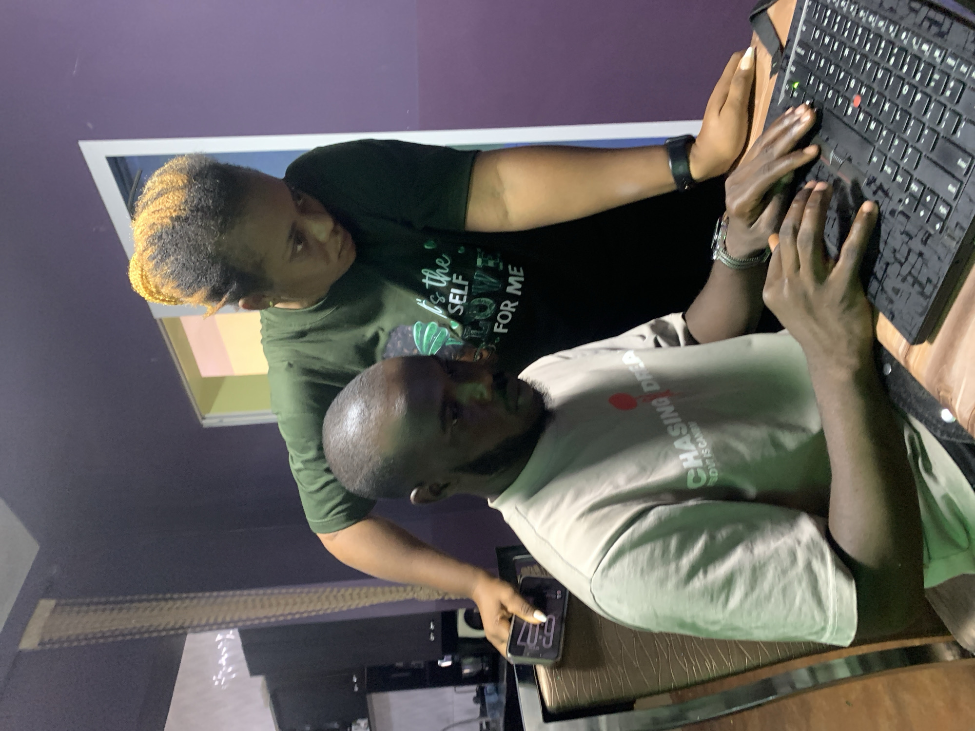 In a residential or office setting within Jos, Nigeria, a male is seated at a table, focused on a laptop, while a female stands leaning over his shoulder, observing the screen.
The seated male has a dark skin tone, short dark hair, and a short beard. He wears a light beige/tan short-sleeved t-shirt with the partially visible text "CHASING DREA" and "LOVE IS CANDI" on the front. A silver-colored watch or bracelet is on his left wrist. His hands are positioned over a black laptop keyboard, appearing to be typing or manipulating the keys. His gaze is directed downwards at the laptop.
The standing female has a dark skin tone and her hair is styled with prominent orange/blonde streaks mixed with dark hair, pulled up. She wears a dark green short-sleeved t-shirt with the full phrase "IT'S THE SELF LOVE FOR ME" printed in light green on the front. A dark-colored smartwatch or fitness tracker is on her left wrist. Her left hand, with long, light-colored fingernails, rests on the table surface next to the laptop. Her right hand is positioned behind the seated male, holding a dark-colored smartphone against the back of his chair. The smartphone screen, visible to the viewer, displays "6:17" and a charging icon. She is leaning forward, looking down at the laptop screen.
The laptop is black, with a visible red trackpoint on the keyboard. It sits on a light-colored wooden table. The seated male occupies a brown chair with a textured, patterned back and seat. The room features predominantly purple walls. To the far left in the background, dark cabinets and what appears to be a kitchen sink or counter area are visible, along with a vertical, light brown, textured blind or curtain. To the right, a white doorframe reveals a section of wall that is light yellow and possibly pinkish.