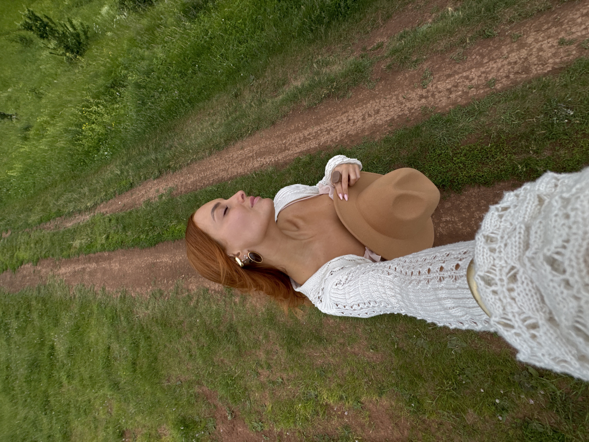 A woman with red hair is sitting on a dirt path in a grassy field. She is wearing a white knit sweater and holding a tan hat. Her eyes are closed, and she appears to be relaxed. The path is surrounded by lush green grass, and trees can be seen in the background. The location is the village of Olkhovka, Russia.