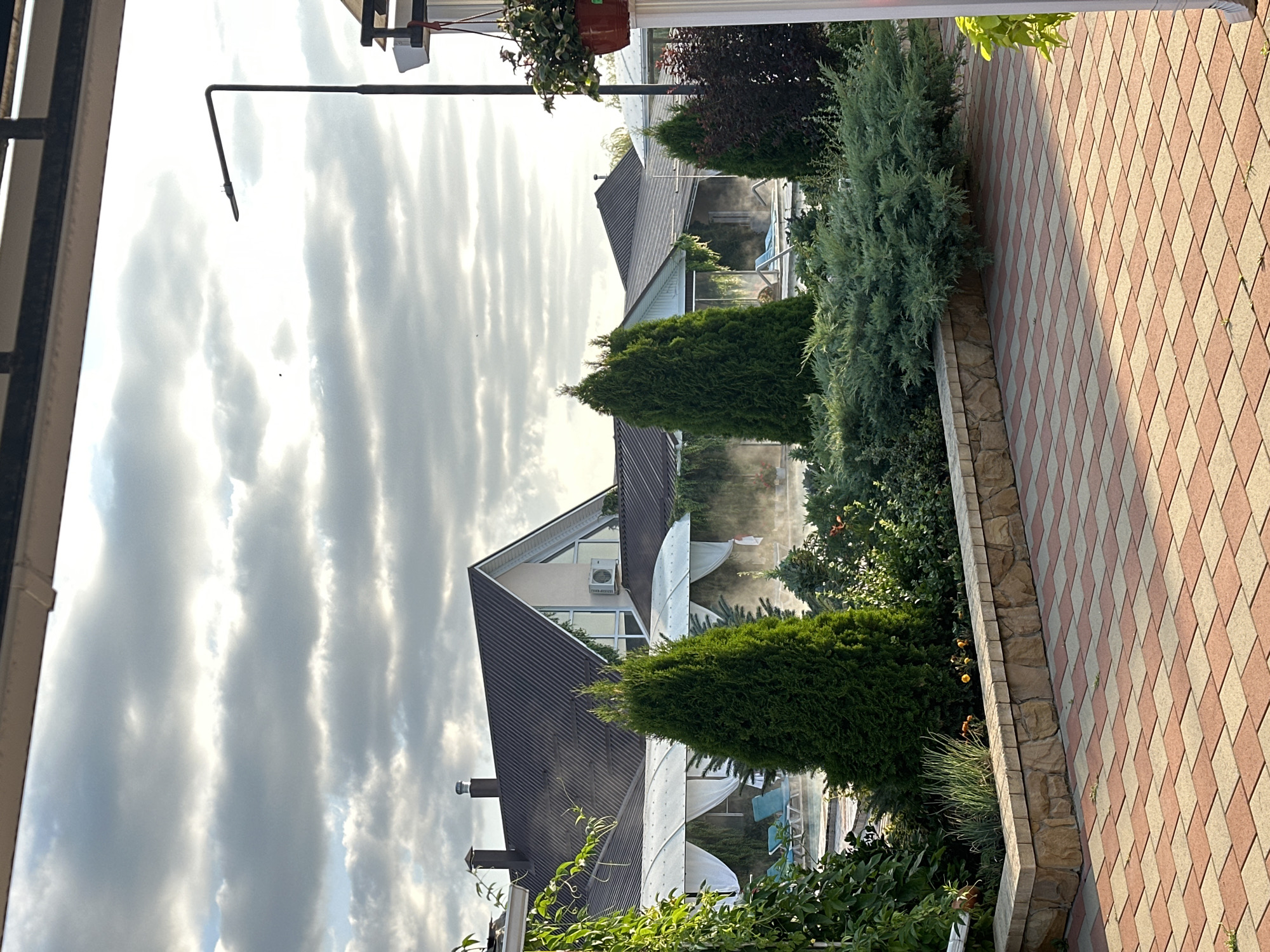 The image depicts an outdoor area with a swimming pool and lounging chairs under white canopies. There is lush greenery, including several evergreen trees and shrubs, in the foreground. In the background, there are modern houses with dark roofs. The sky is partly cloudy, with sunlight filtering through. The paving in the foreground is made of reddish-brown and beige rectangular tiles arranged in a diagonal pattern. The overall setting suggests a resort or a residential area with recreational facilities. The exact location in Yaroslavskaya, Russia, is not identifiable from the visual elements alone.