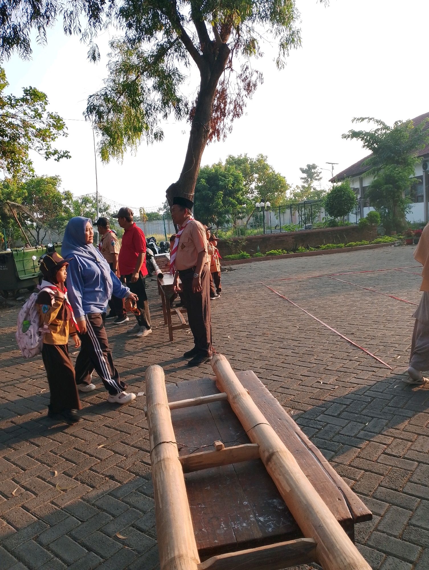 The image depicts an outdoor scene in Probolinggo, Indonesia, possibly a schoolyard or public park, characterized by a paved courtyard with interlocking bricks. The time of day is likely late afternoon, judging by the long shadows cast by the people and objects. In the foreground, a makeshift wooden structure made of logs and planks is prominently displayed.
Several individuals are present in the image, many of whom appear to be participating in a scouting or similar youth activity, indicated by their uniforms. On the left, a young person wearing a brown uniform and backpack is walking alongside an adult in a blue headscarf and dark pants. To their right, an adult male in a red shirt and cap is standing and interacting with another individual. Further to the right, a man in a scout uniform with a sash is engaged in conversation, facing towards the left. In the background, more people in similar uniforms are visible, suggesting a group event or gathering.
The environment includes lush green trees and foliage, a building with a tiled roof in the mid-ground, and a fence. The overall atmosphere appears to be casual and communal, with people interacting and engaging in activities outdoors. The bright sunlight and clear sky indicate favorable weather conditions. The paving stones are laid in a herringbone pattern, common in outdoor public spaces. A red line is marked on the pavement, potentially for a game or activity.