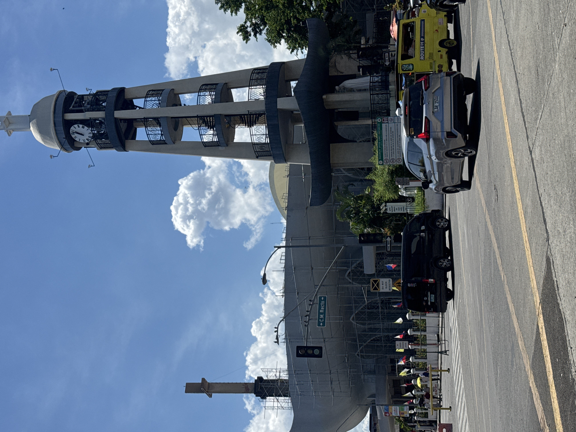 The image depicts a street scene in Davao City, Philippines, with a focus on the San Pedro Cathedral under construction. A tall, modern clock tower stands prominently on the right, adorned with a clock face showing Roman numerals. The cathedral, on the left, is partially covered in scaffolding, suggesting ongoing renovations or expansion. Several vehicles, including a black van, a silver SUV, and a yellow jeepney, are visible on the road, indicating a busy urban environment. Pedestrians can be seen on the sidewalk, some holding umbrellas. Traffic signals and street signs are present, further emphasizing the city setting. The sky is a clear blue with scattered white clouds, suggesting fair weather.