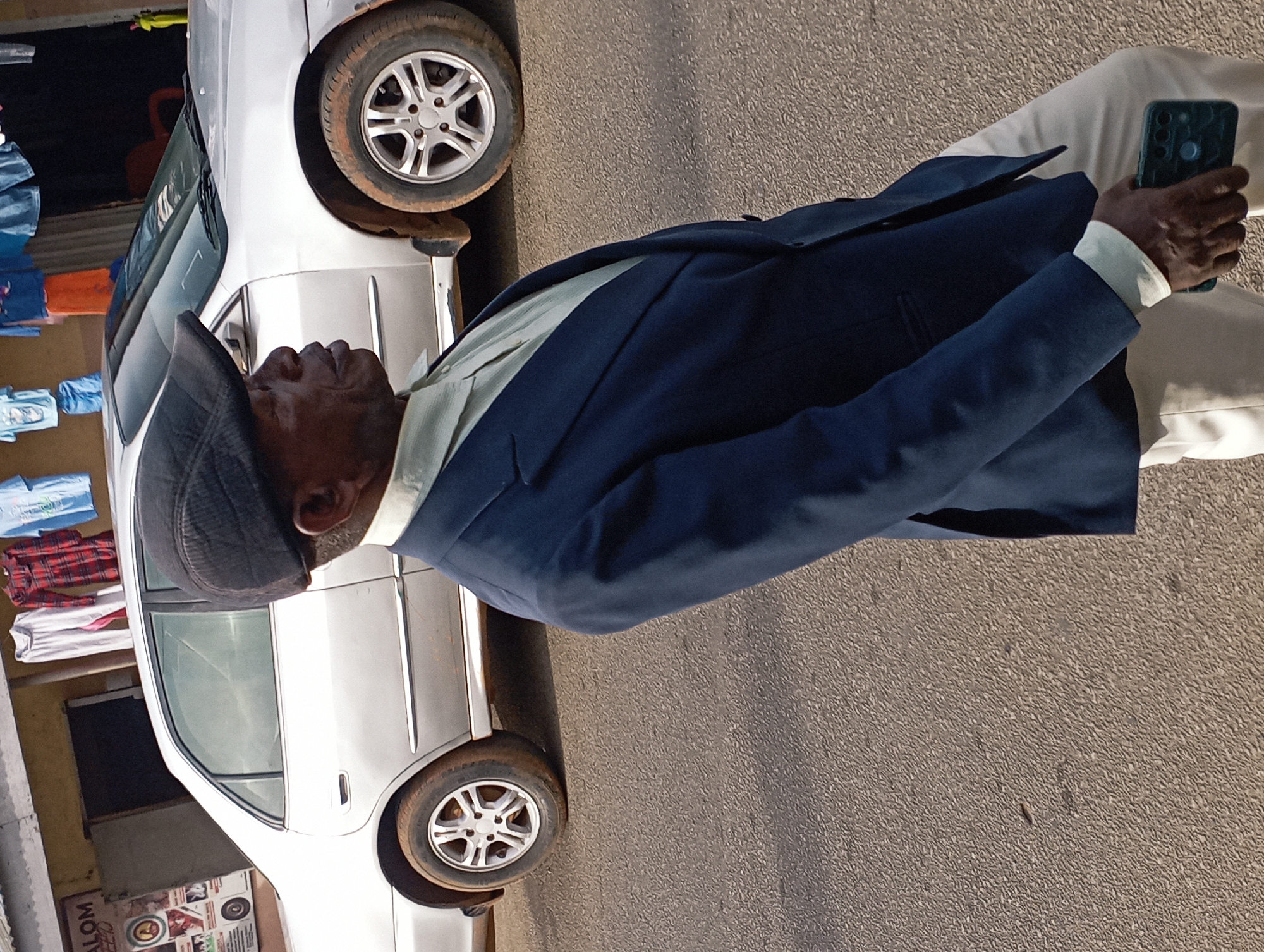 A man in a dark blue suit jacket and cream-colored pants walks on a paved street. He is wearing a dark flat cap and a light-colored collared shirt. He is holding a dark green smartphone in his right hand. Behind him, a silver sedan is parked on the side of the road. Clothes are displayed for sale on a rack in front of a building behind the car. The scene appears to be in Jos, Nigeria, indicated by a sign in the background that reads "SALOM SEED." The paved street has a textured surface.