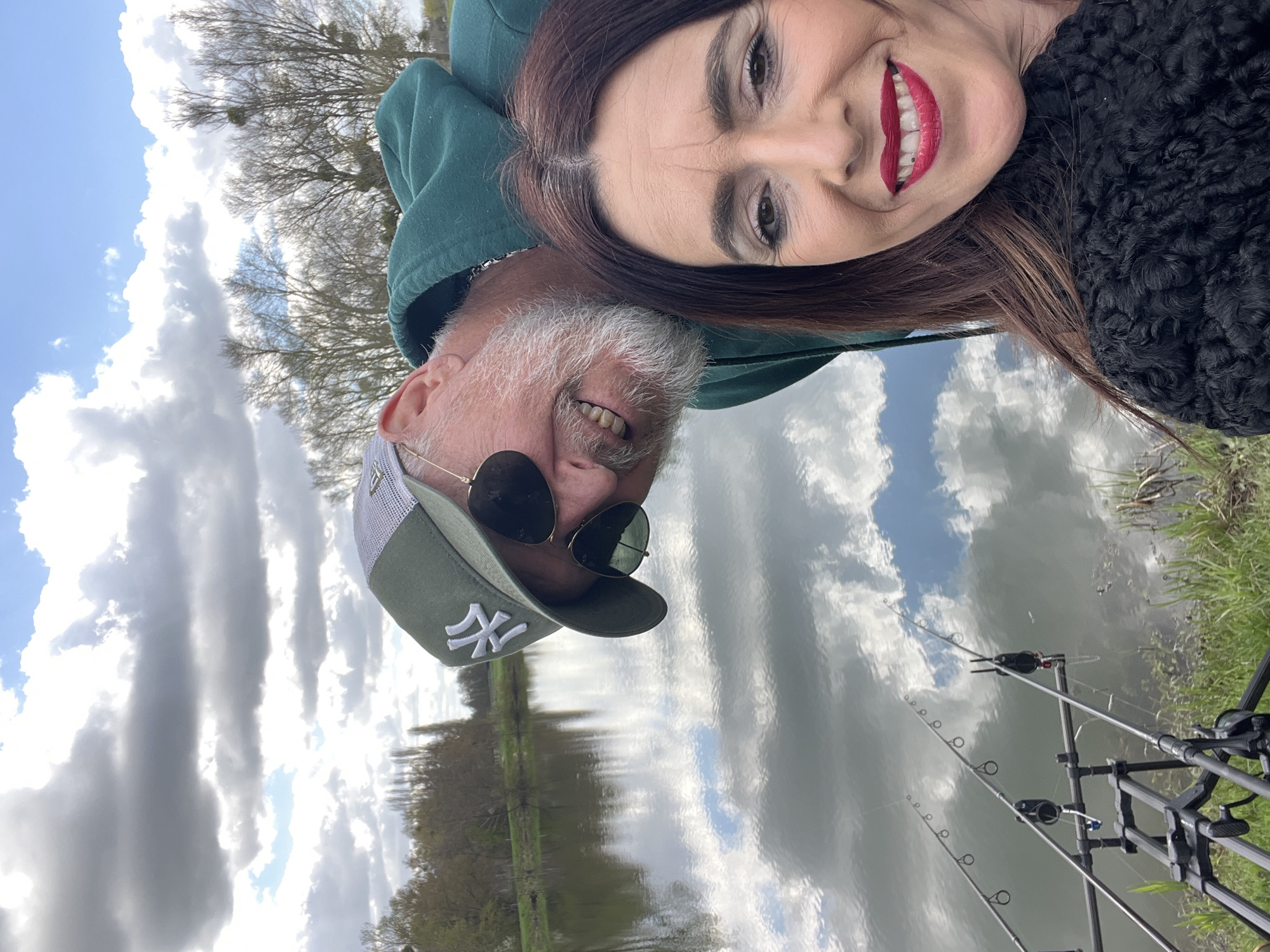 A selfie photograph taken outdoors depicts two people, likely a couple, in the context of a fishing activity. The woman is dark-haired and wearing red lipstick, while the man has a gray beard, sunglasses, and a green baseball cap with a white symbol. The photo appears to be taken at a slight angle, resulting in a tilted perspective. In the background is a body of water reflecting the sky with clouds. Fishing rods and equipment are visible near the water's edge. The location is Gavisse, France.