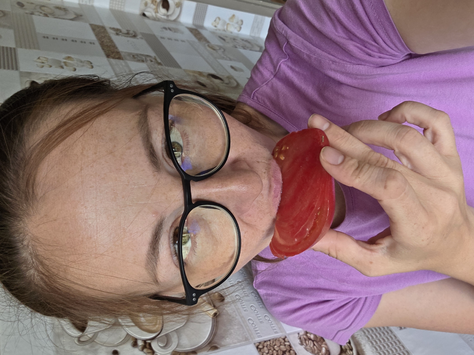 A woman wearing glasses and a purple t-shirt is holding a slice of tomato to her mouth, as if to eat it. Her hair is pulled back, and she has freckles on her face. The background appears to be a kitchen or dining area, with a patterned wallpaper. The image was taken in Chaikovsky, Russia.