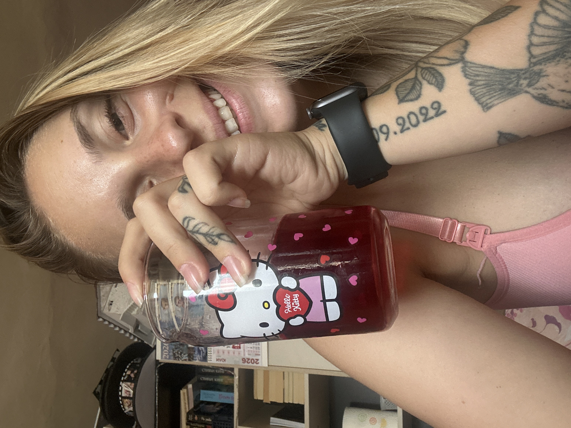A woman with blonde hair and freckles holds a glass of red liquid with Hello Kitty design. She is wearing a pink bra. A tattoo of dates "09.2022" is visible on her arm, along with other tattoos of nature motifs. She is in an indoor setting with bookshelves in the background. The image is a selfie taken from a low angle. The location is not Plovdiv, Bulgaria.