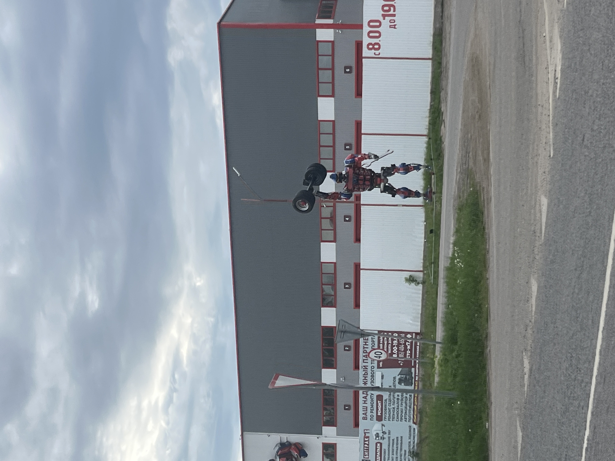 The image shows a roadside scene in Staraya Kupavna, Russia. A sculpture of a robot figure made from scrap metal stands near a gray building with red trim and a white section with red text. A sign with Cyrillic text related to vehicle repair is visible. The road itself is asphalt, and the sky is cloudy.