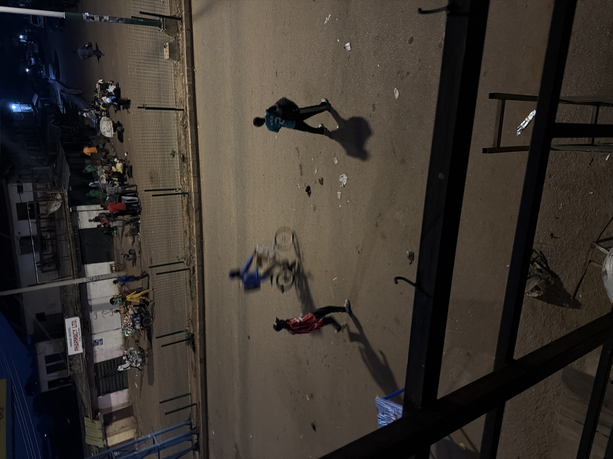 In Bukuru, Nigeria, the image captures a street scene at dusk. A busy intersection is visible, with people on foot and a cyclist in motion. The cyclist, wearing a blue shirt, is a blur of motion on a bicycle. Another individual in a red jacket and black pants walks across the street, casting a long shadow. A third person, wearing a teal jersey with the number 2, walks away from the camera, carrying a large dark bag over their shoulder. In the background, a row of shops and buildings lines the street, some illuminated by artificial lights. Pedestrians and small groups of people are gathered near the shops. A cart laden with goods is also visible, with several individuals around it. The street is paved with asphalt and shows signs of wear, with scattered litter. The overall impression is of a lively, active street scene in an urban environment transitioning from day to night.
