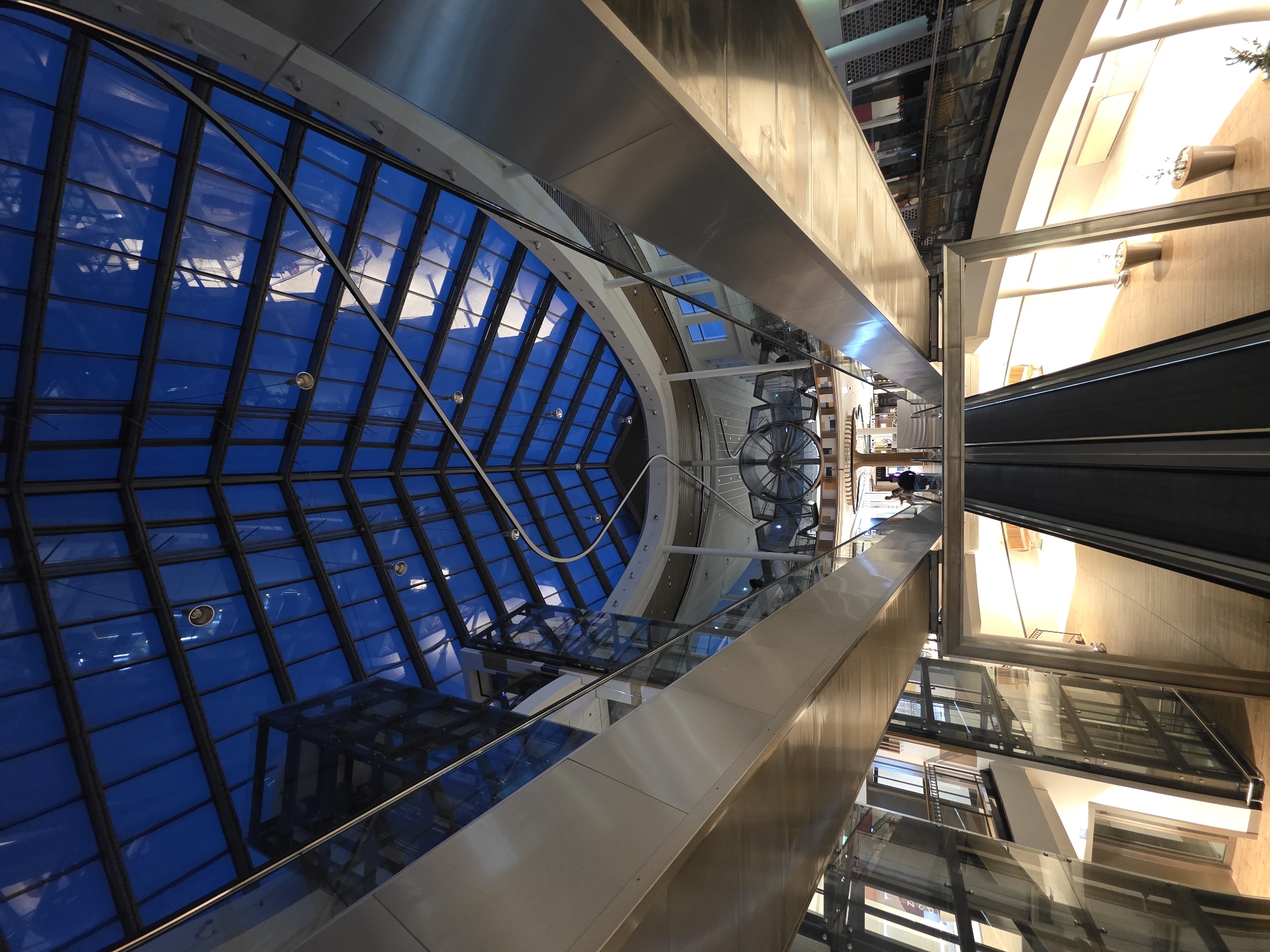 This is a low-angle, wide shot of the interior of a modern shopping mall with a large, multi-faceted glass and metal atrium roof. The roof is dark blue and reflects the lights of the interior. The mall features multiple levels with walkways, escalators, and glass elevators. The architecture is sleek and contemporary, with polished metal railings and wooden flooring. In the foreground, two escalators are visible, leading upwards. On the walkways, a few people are seen in the distance, including a person with a stroller. The lighting is a mix of natural light filtering through the roof and artificial light from within the mall. The overall impression is one of spaciousness and modern design.